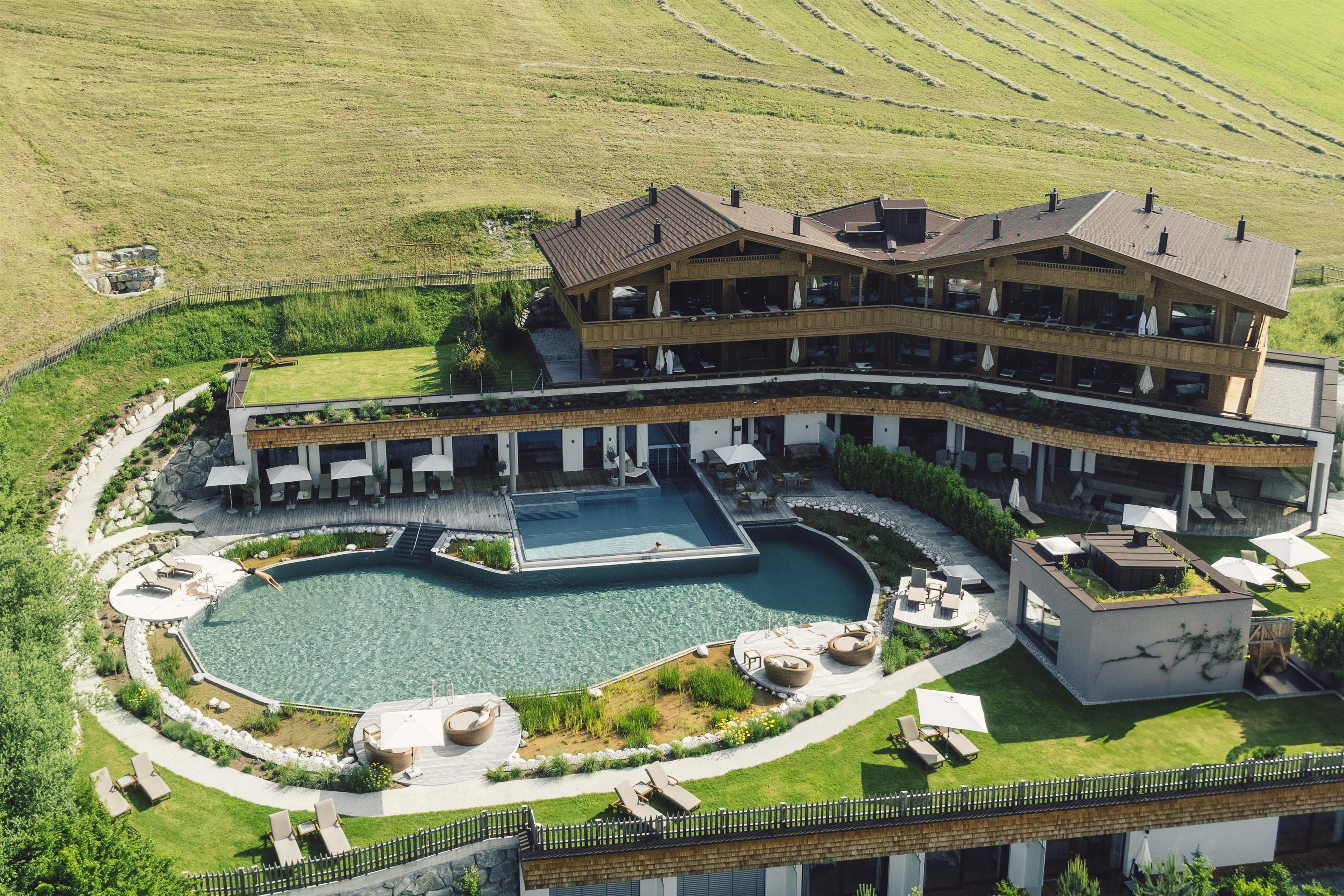 A modern hotel with multiple floors, surrounded by green grounds. In the foreground, there is an elegant swimming pool with loungers and a beautiful garden layout.