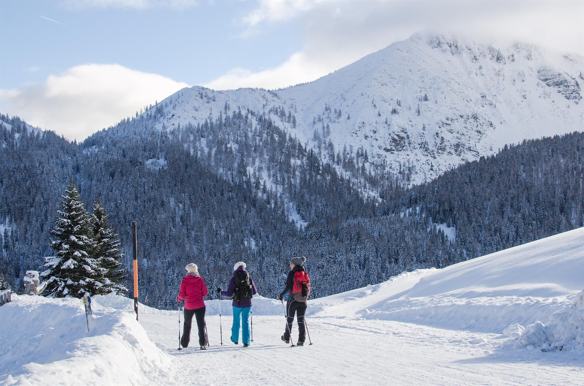 Three people are standing on a snowy path, looking at snow-covered mountains. The sun is shining and the sky is partly cloudy.