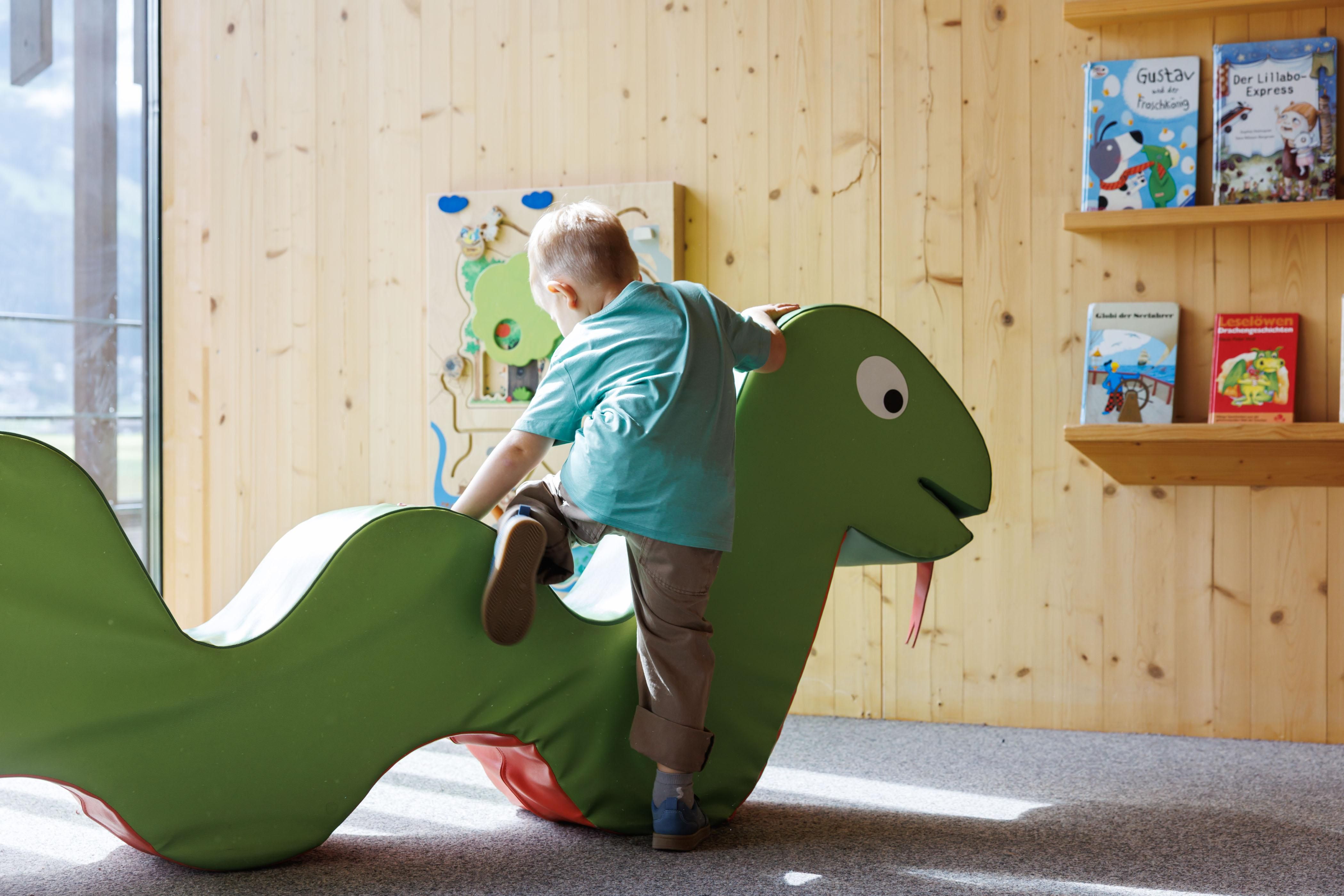 A child is playing on a green snake figure in a bright room. In the background, there are shelves with children's books.
