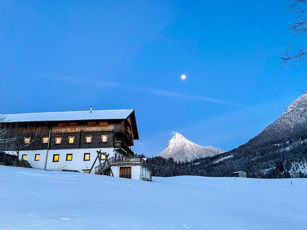 A picturesque mountain cabin in the snow, surrounded by majestic mountains. The clear sky and the moon give the scene a tranquil atmosphere.