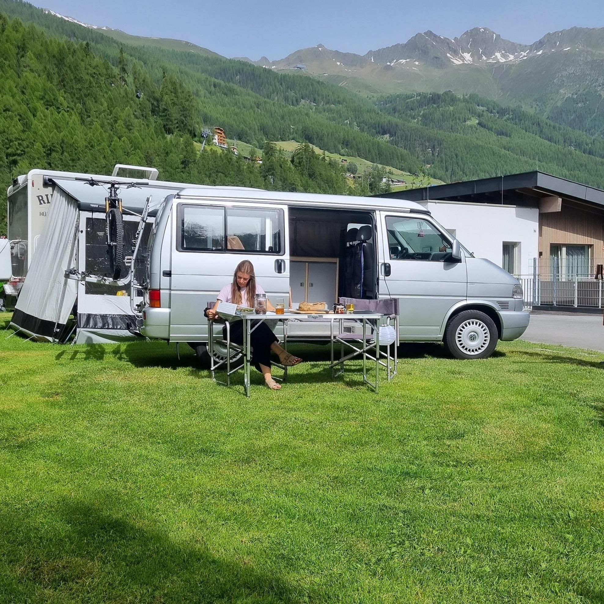 A motorhome is parked on a green meadow with a view of the mountains. A woman is sitting at a table and enjoying a meal outdoors.