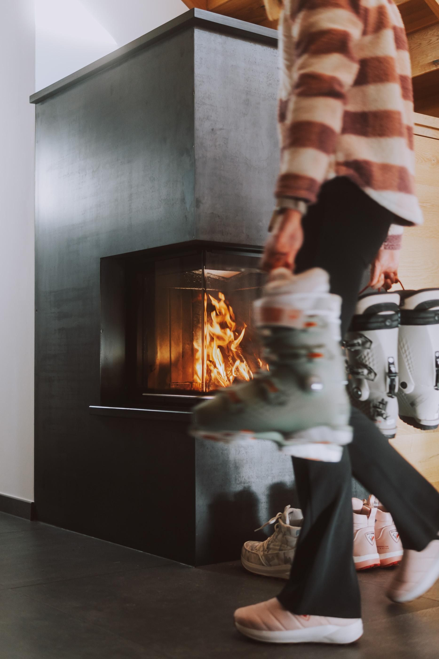 A cozy living space with a modern fireplace. A person is wearing ski or snowboard boots and moving through the room.