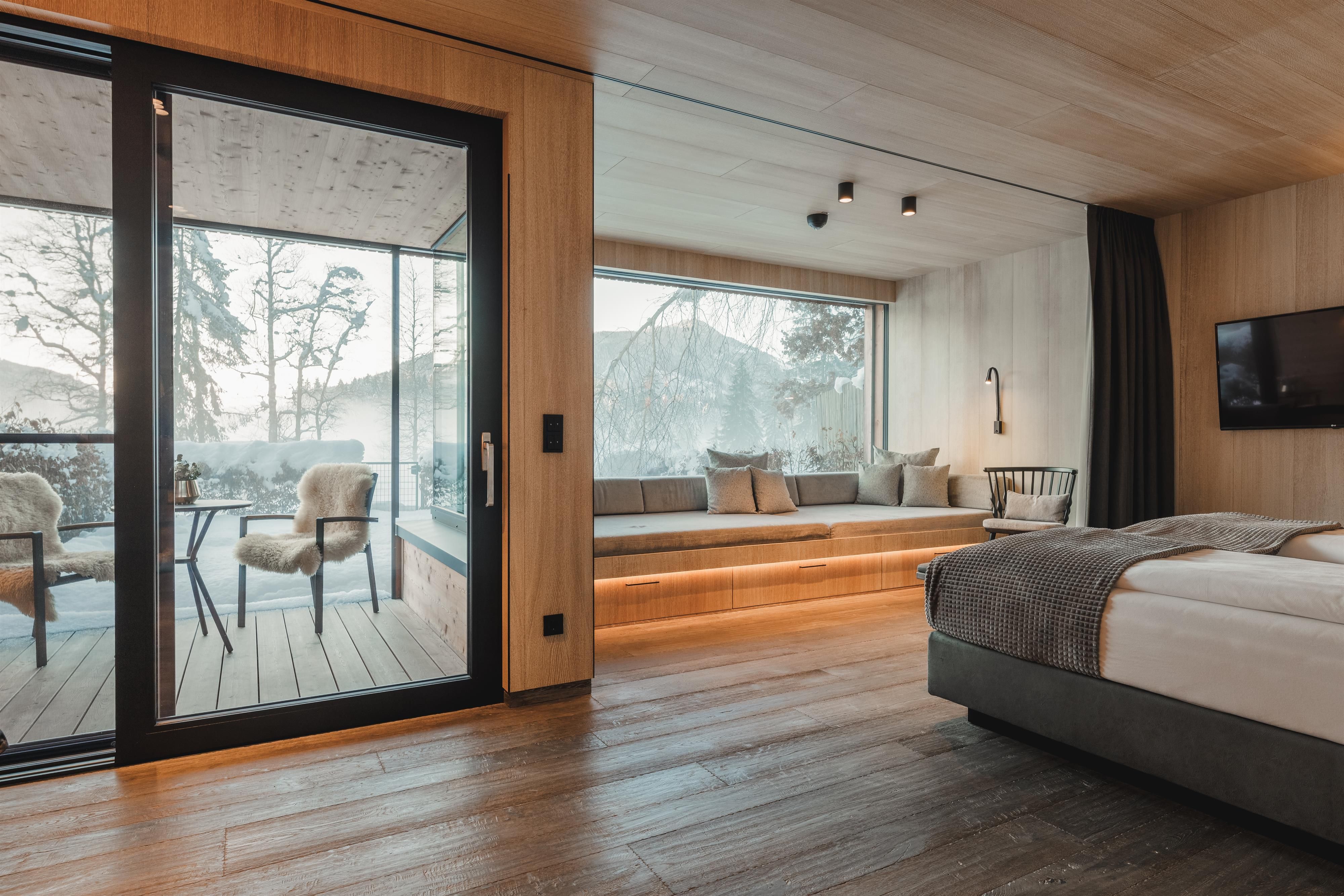 A modern hotel room with a large window and a view of a balcony. The room is furnished with wood and neutral colors, creating an inviting atmosphere.
