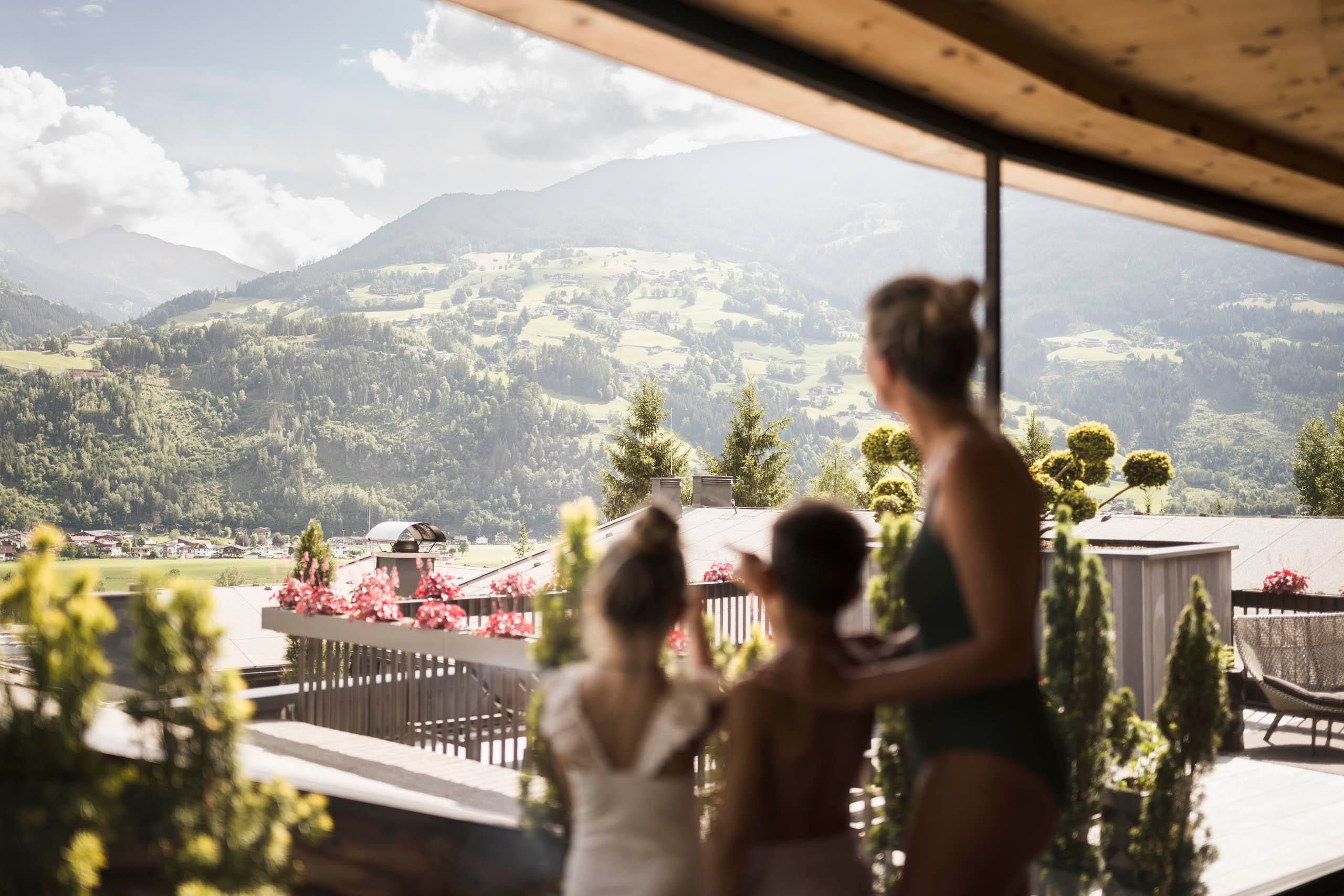 A family stands on a terrace and looks out at a beautiful mountain landscape. The surroundings are adorned with colorful flowers and green plants.