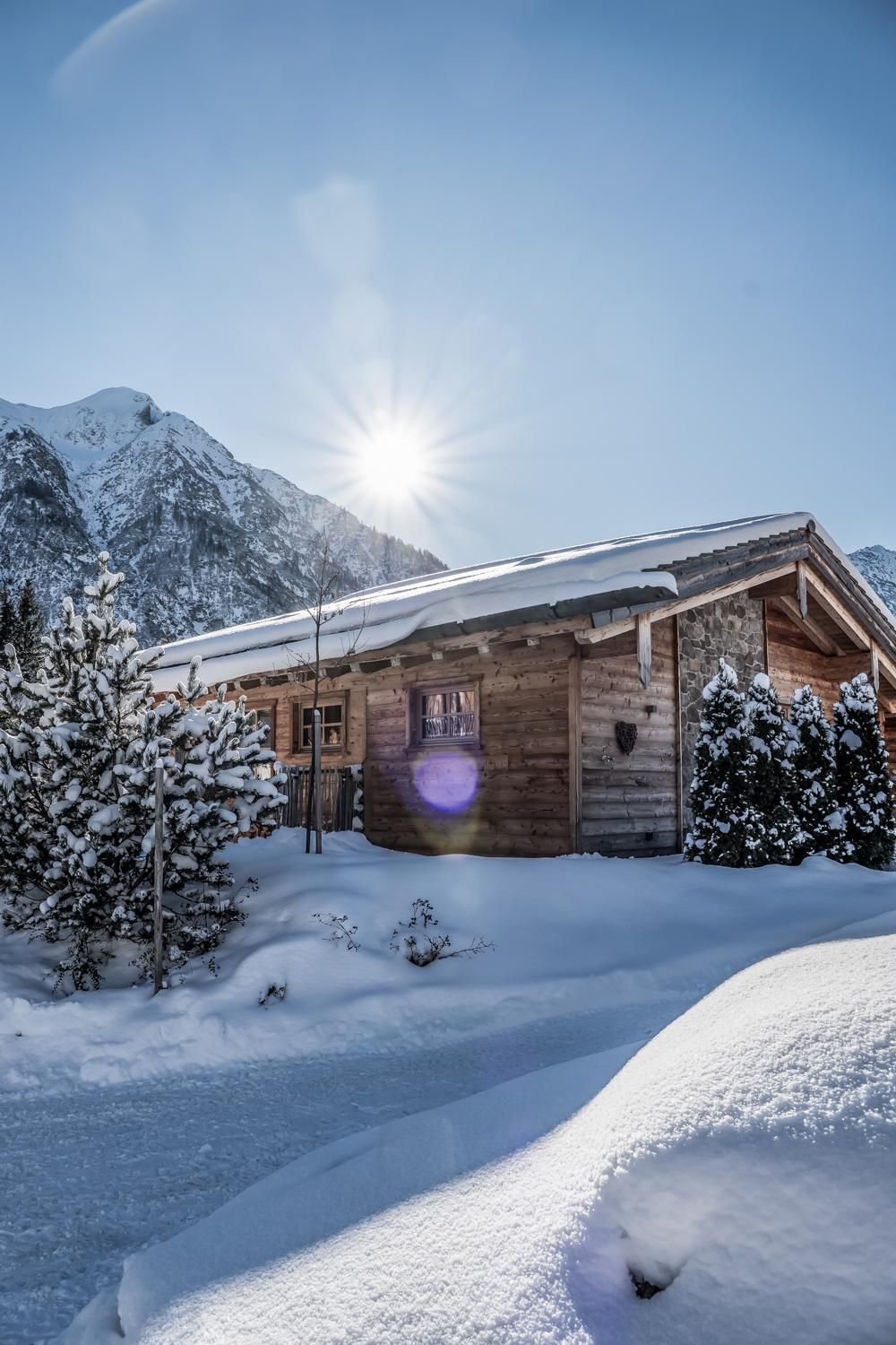 A cozy chalet in the snow, surrounded by snow-covered trees. The sun is shining behind the mountains.