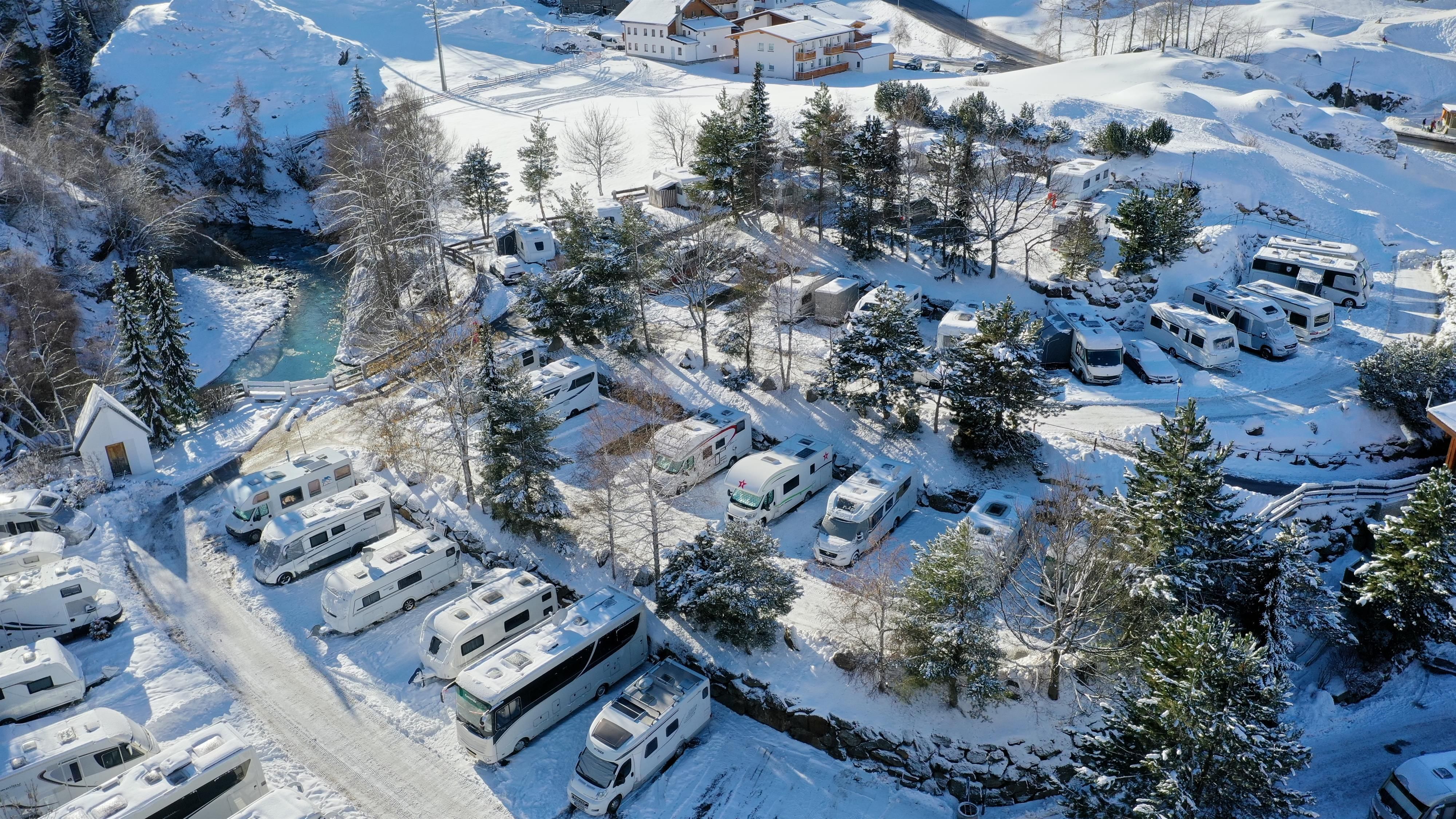 A motorhome park in a snowy landscape. Surrounded by trees and a small river.