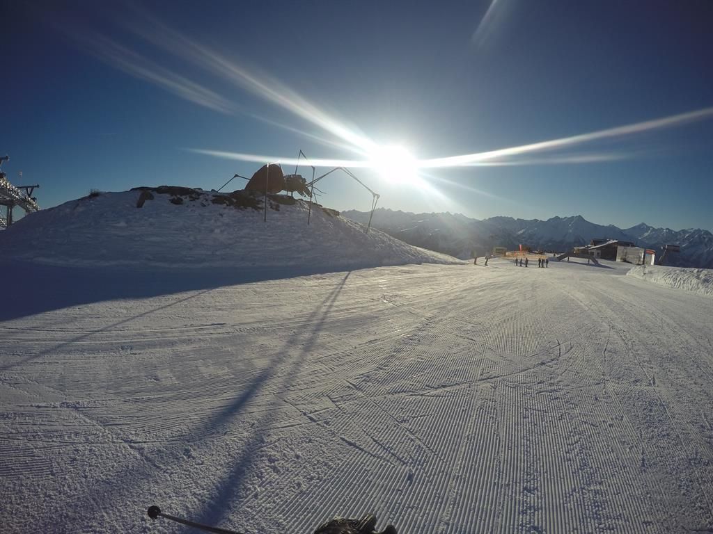 A snow-covered slope under a clear blue sky. The sun shines brightly over the mountains.