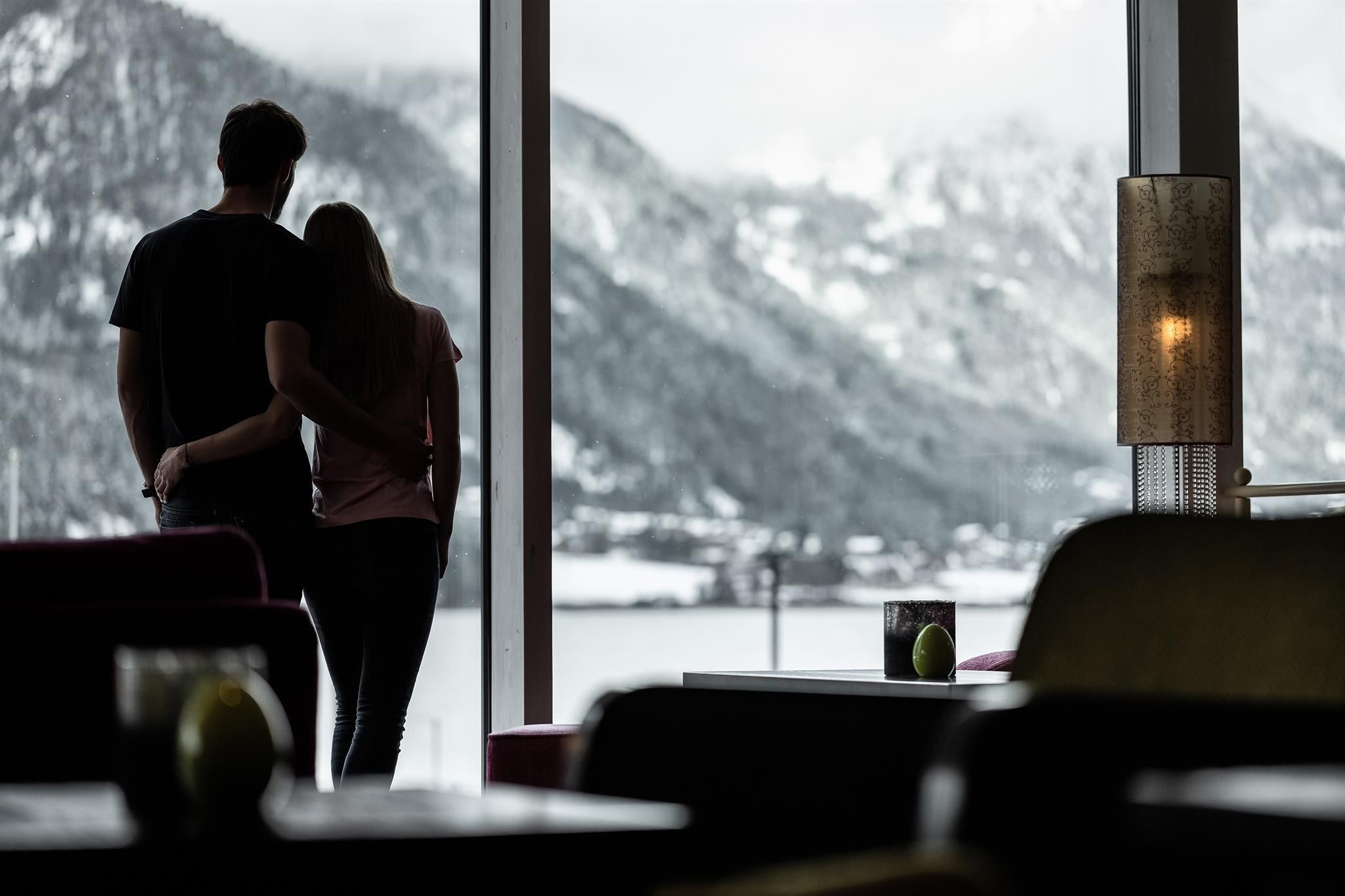 A couple stands by the window and looks out at the snowy mountain landscape. The atmosphere is romantic and cozy.