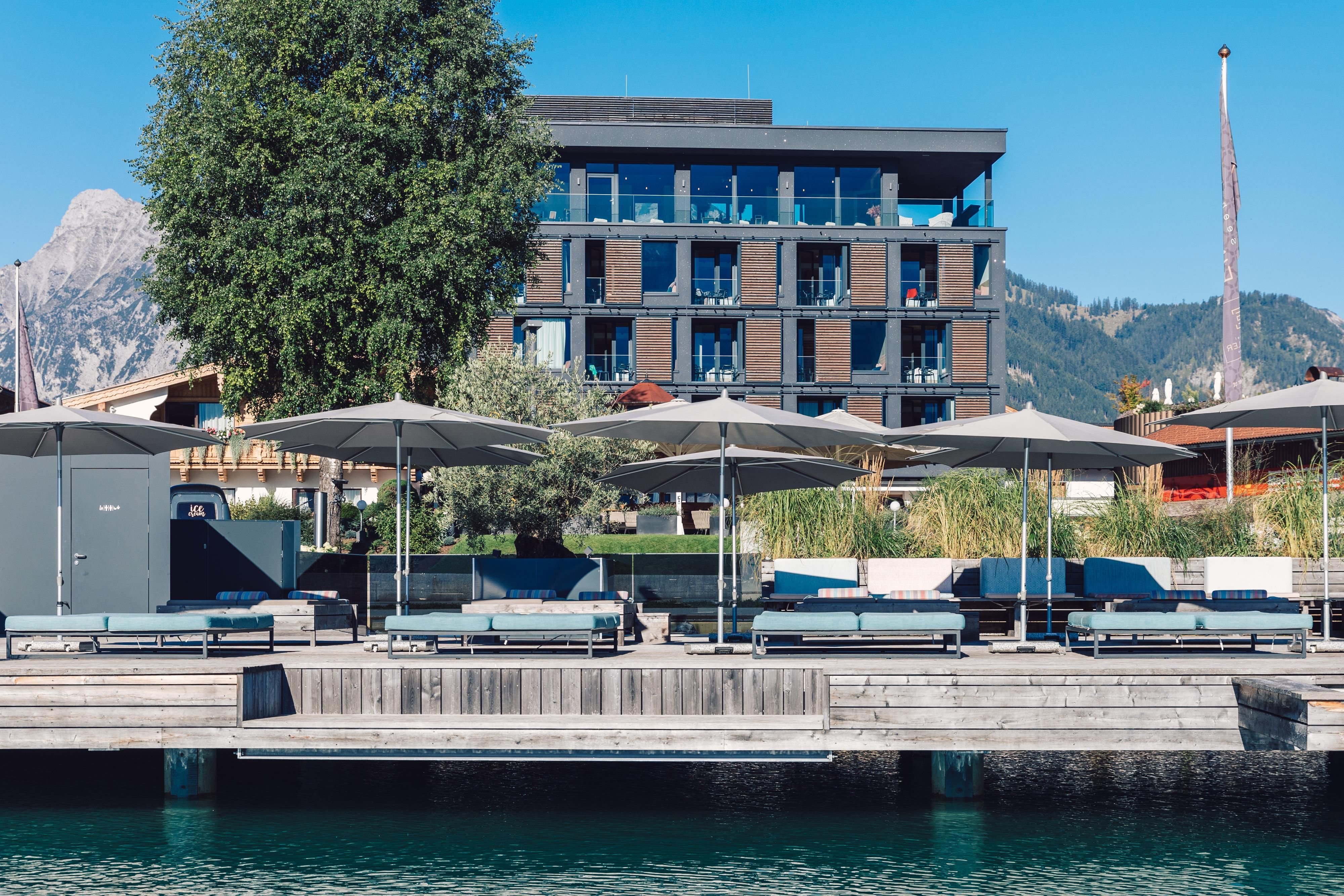 A modern building by the water with sun umbrellas and loungers on the terrace. In the background, mountains and a blue sky are visible.