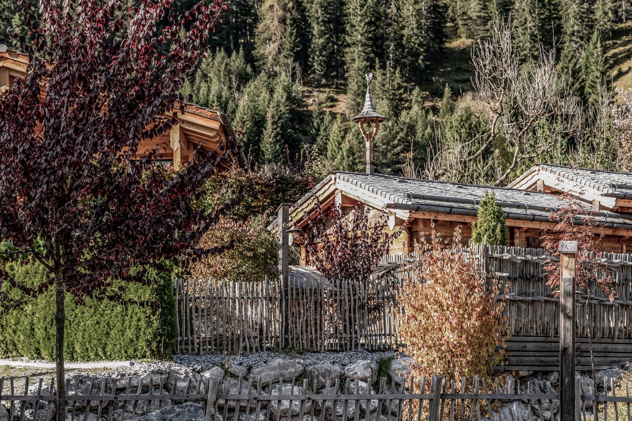 An idyllic landscape with traditional wooden chalets and colorful trees. In the background, gentle hills and dense forest can be seen.