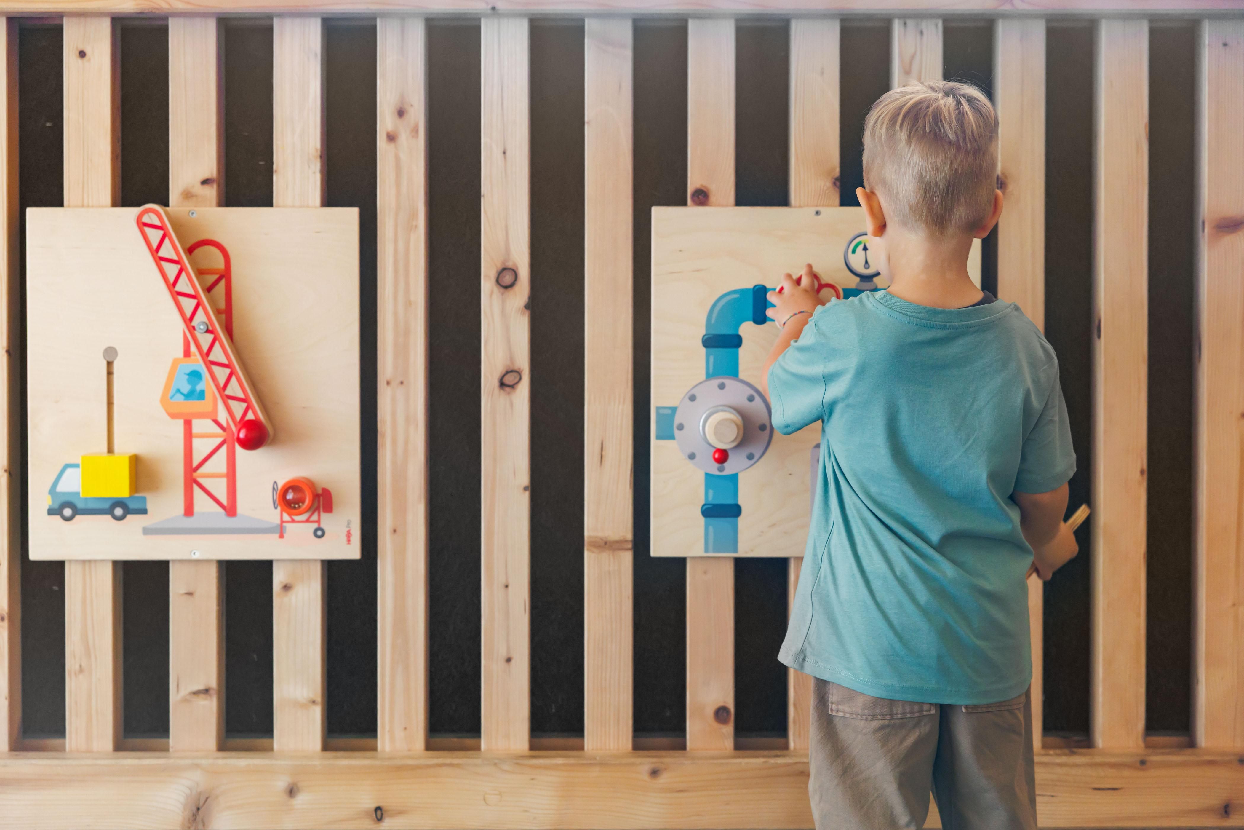A boy stands in front of two wooden panels featuring colorful construction site motifs. He is interacting with a toy crane and a water pipe.