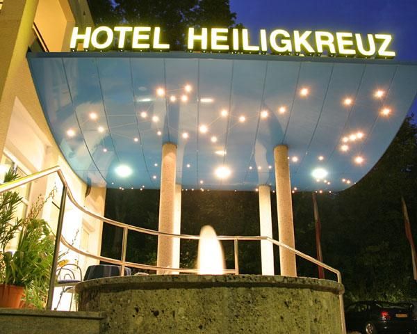 A hotel named "Heiligkreuz" that is brightly lit. The entrance area features a modern canopy and a decorative fountain.