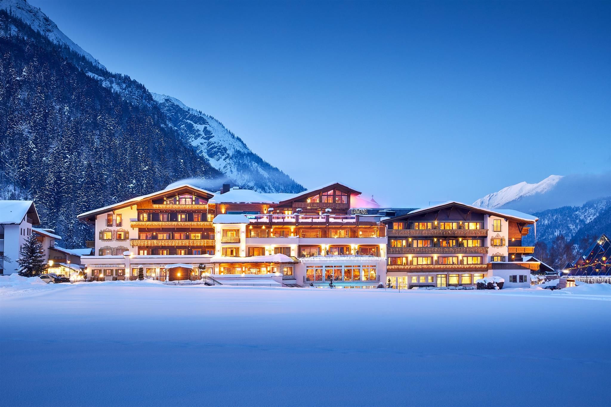 A beautiful hotel in the Alps, surrounded by snow-capped mountains. The façade of the building glows warmly in the evening light.