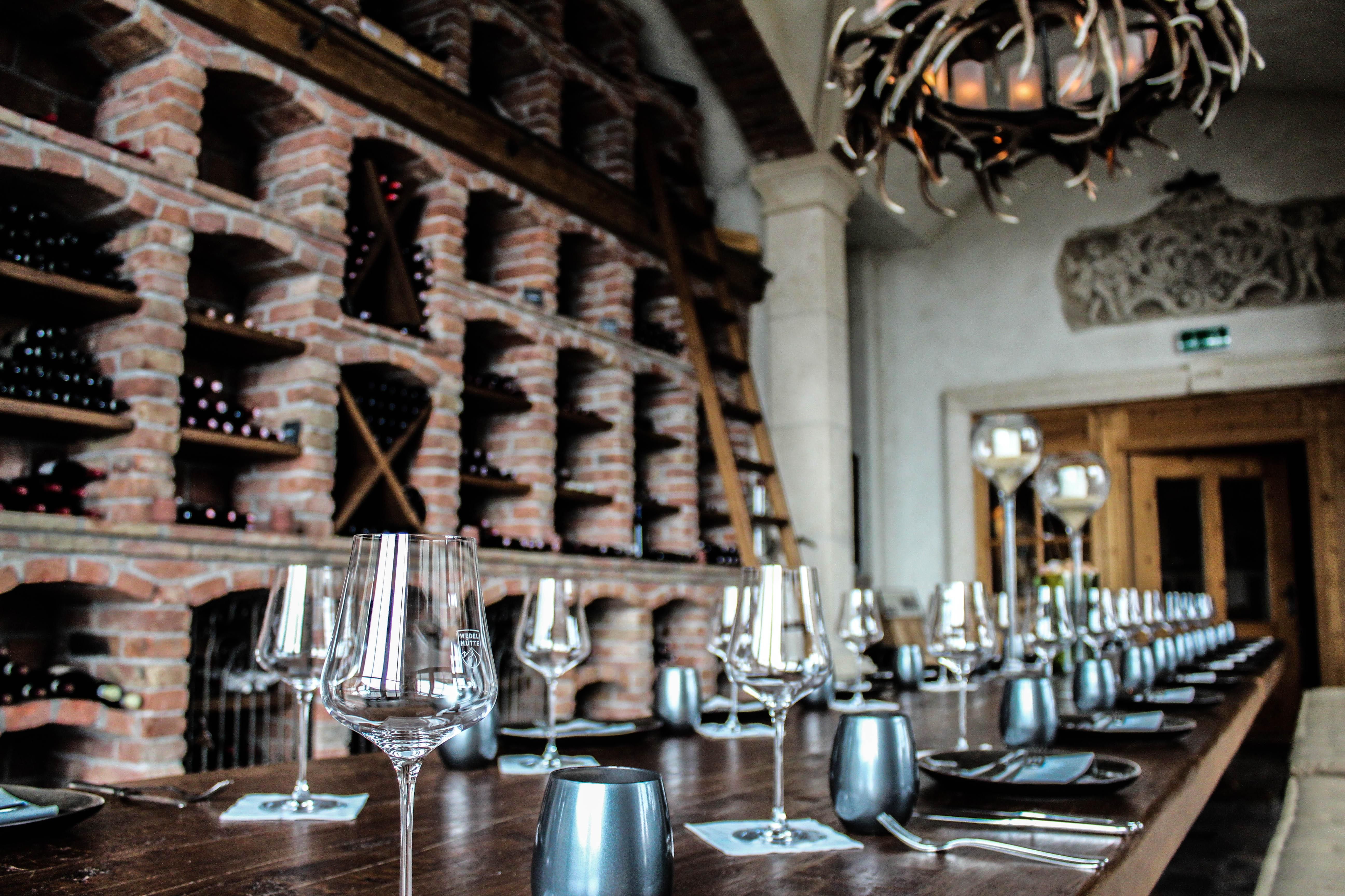 An elegant restaurant with a long wooden table. In the background, there are wine shelves made of brick and an appealing lighting design.