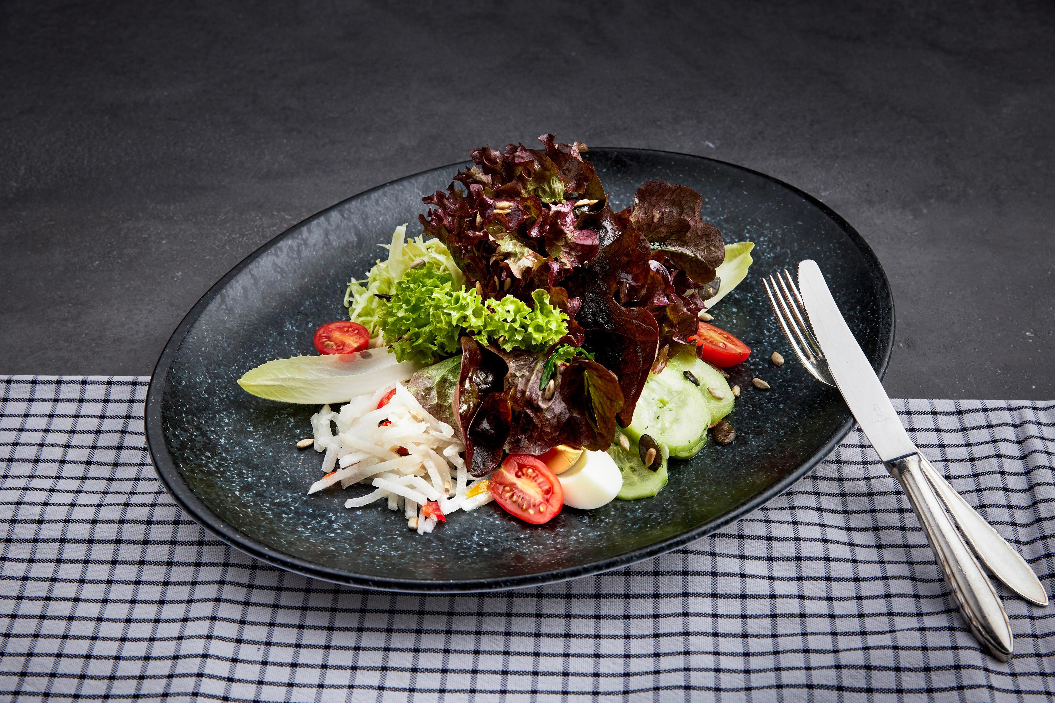 A fresh salad on a black plate, garnished with various lettuce leaves, cherry tomatoes, and cucumbers. The background is simple and elegant.