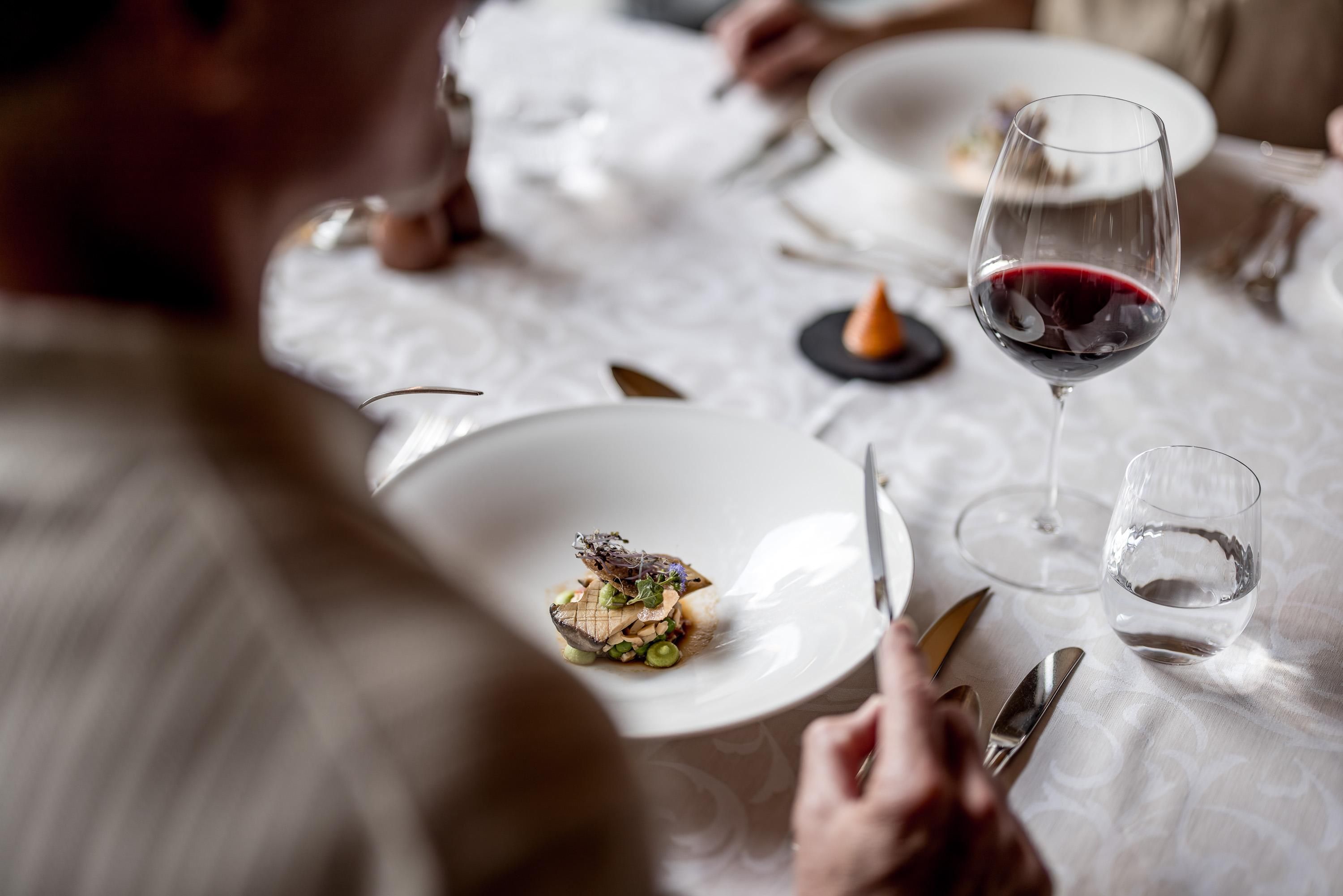 An elegant dish on a table, surrounded by cutlery and glasses. In the background, a glass of red wine is being served.