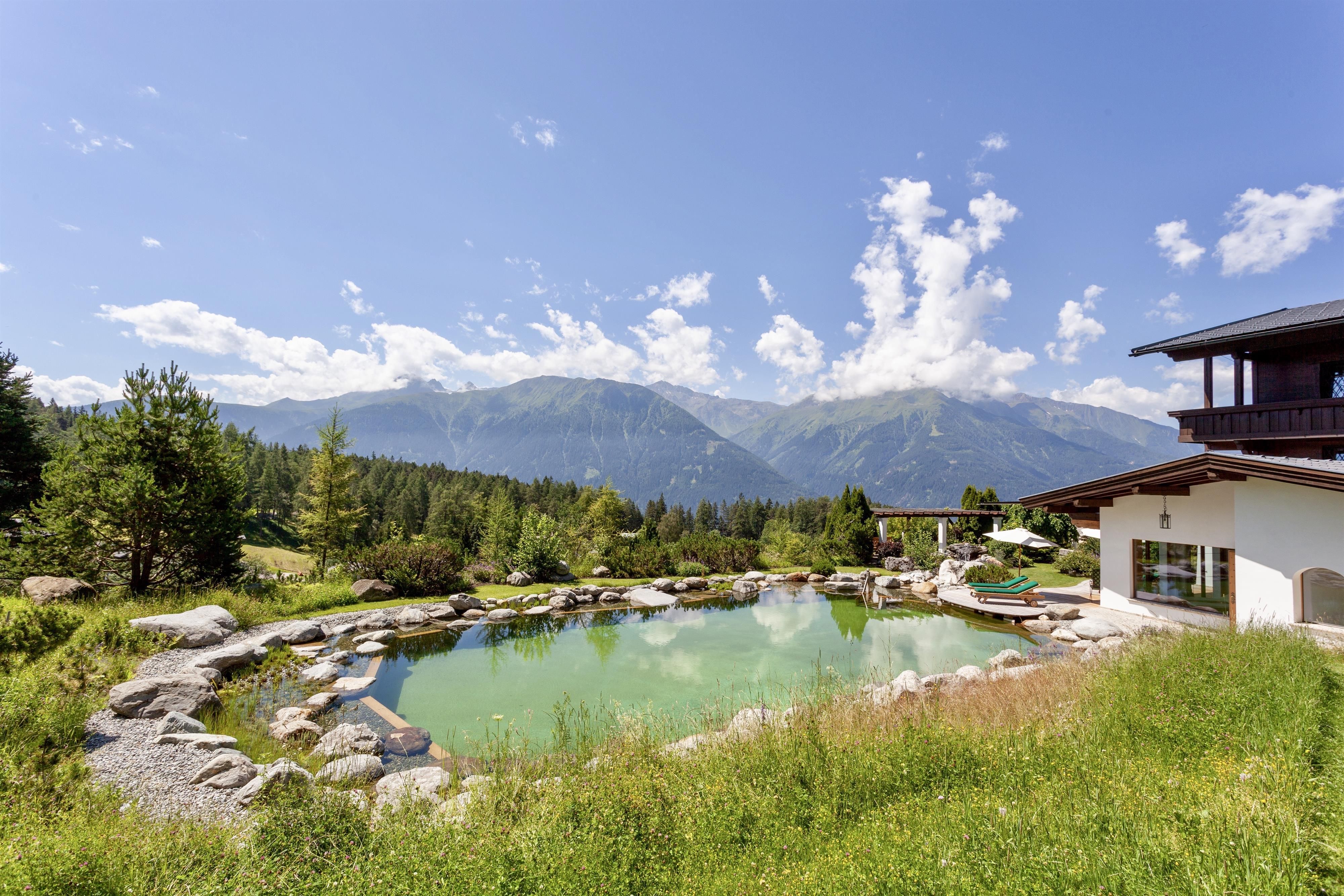 A picturesque pond surrounded by lush greenery and mountains in the background. The sky is clear and sunny, ideal for a relaxing stay in nature.