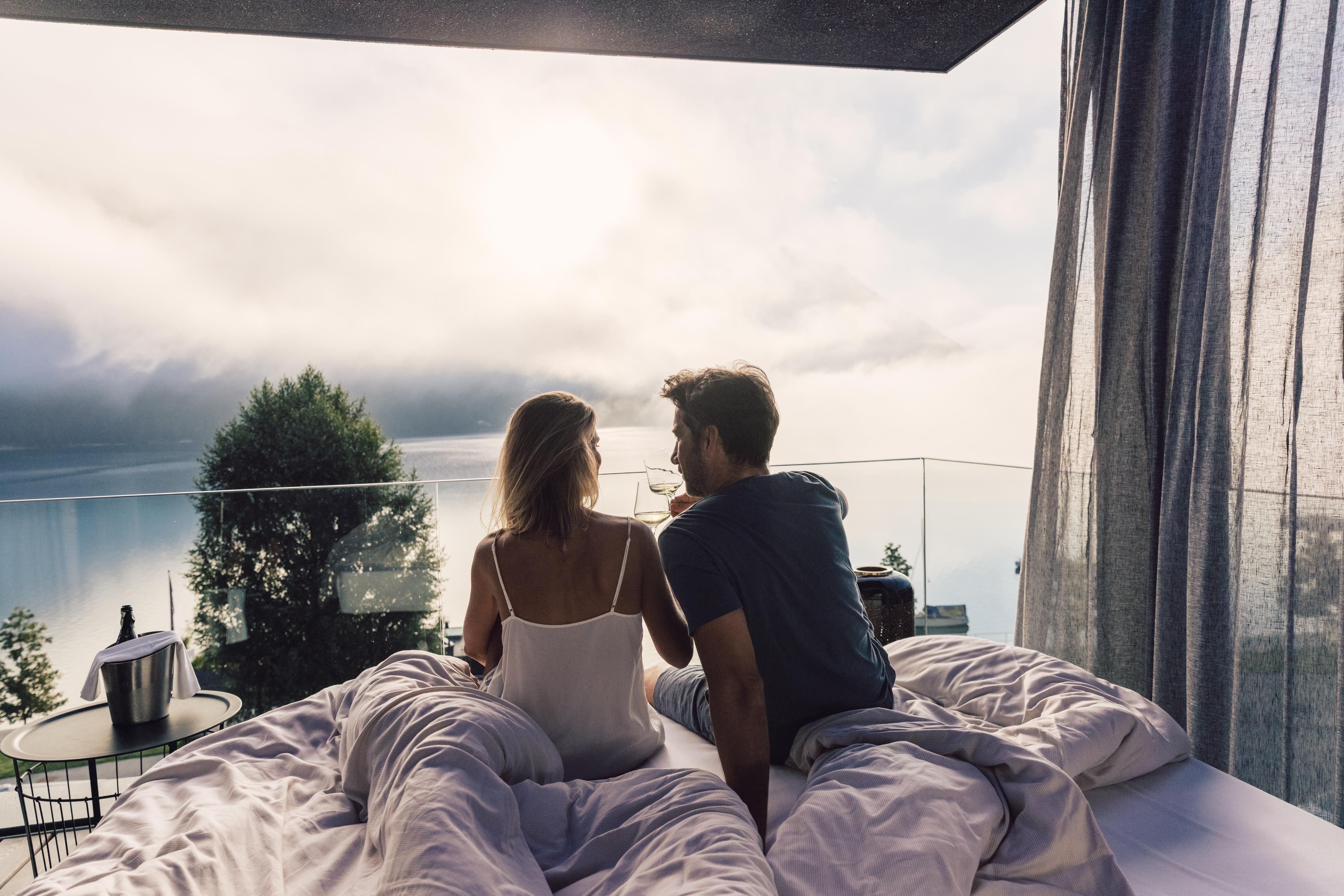 A young couple sits on a bed overlooking a picturesque landscape. They are enjoying a quiet moment with a beautiful view.
