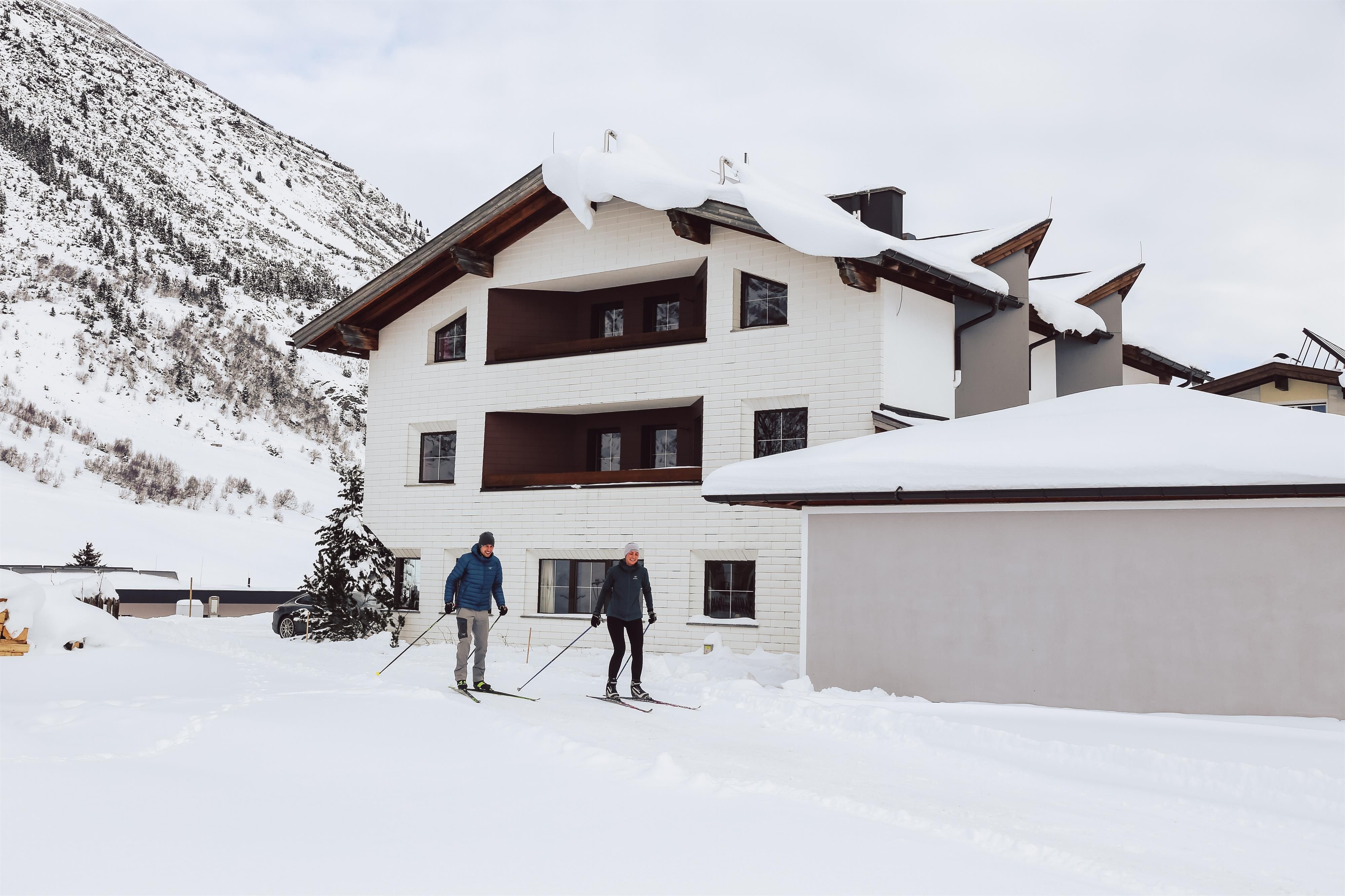 Two skiers stand in front of a white house in the snow. In the background, snow-covered mountains can be seen.