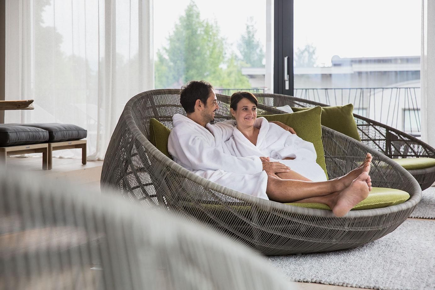 A couple in bathrobes sits relaxed in a modern, woven armchair. They enjoy the serene atmosphere of a bright, elegant room.