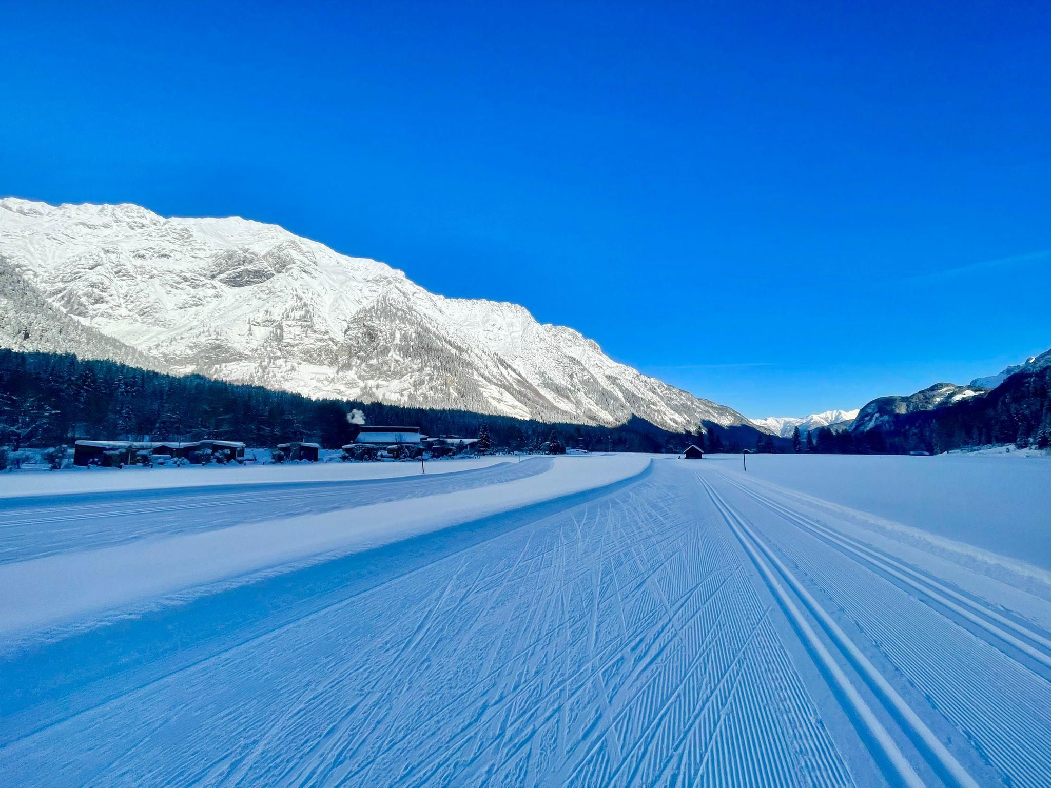 A snow-covered landscape with mountains and a clear blue sky. The slope runs through the impressive winter scenery.
