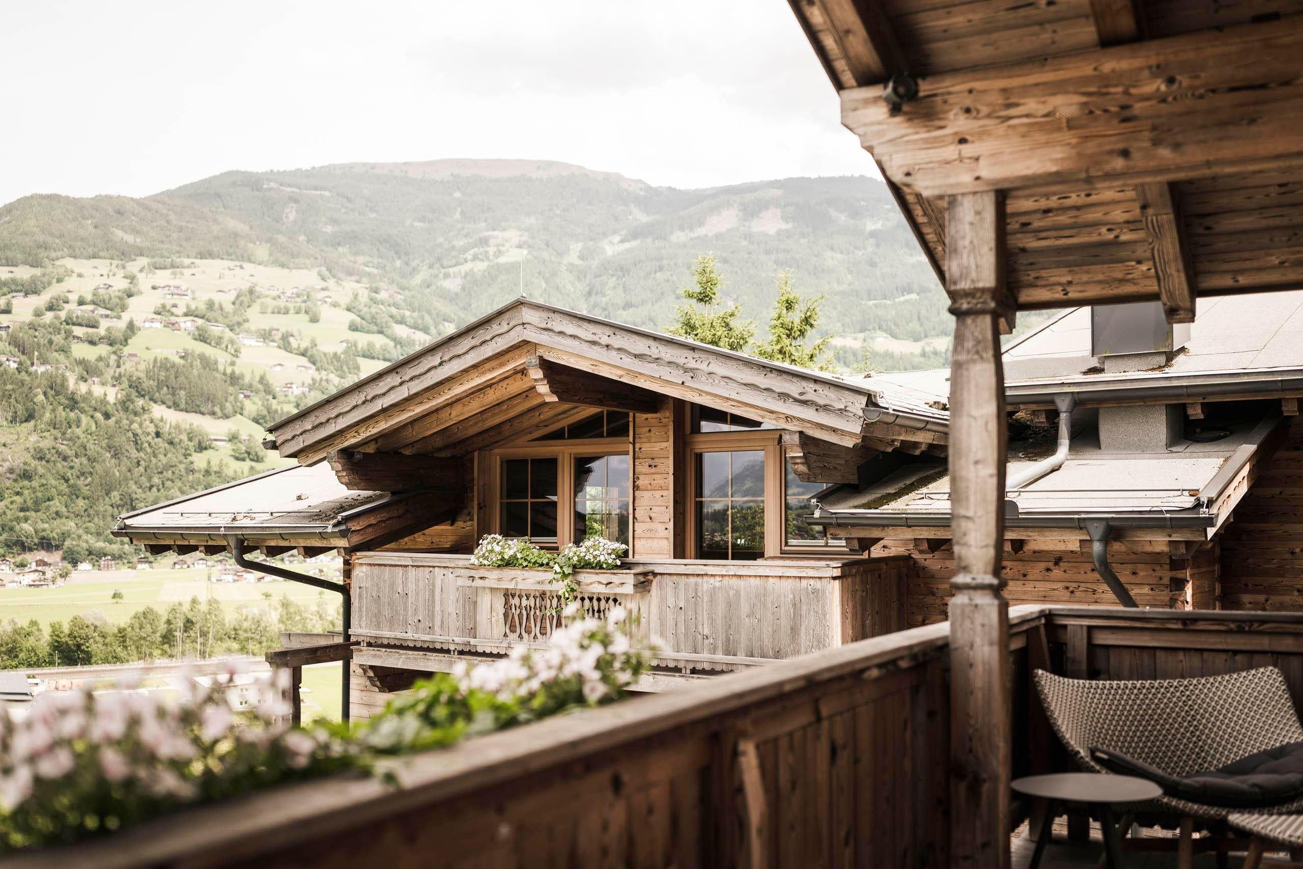 A cozy wooden house with a balcony and a beautiful view of the mountains. The surroundings are green and inviting.
