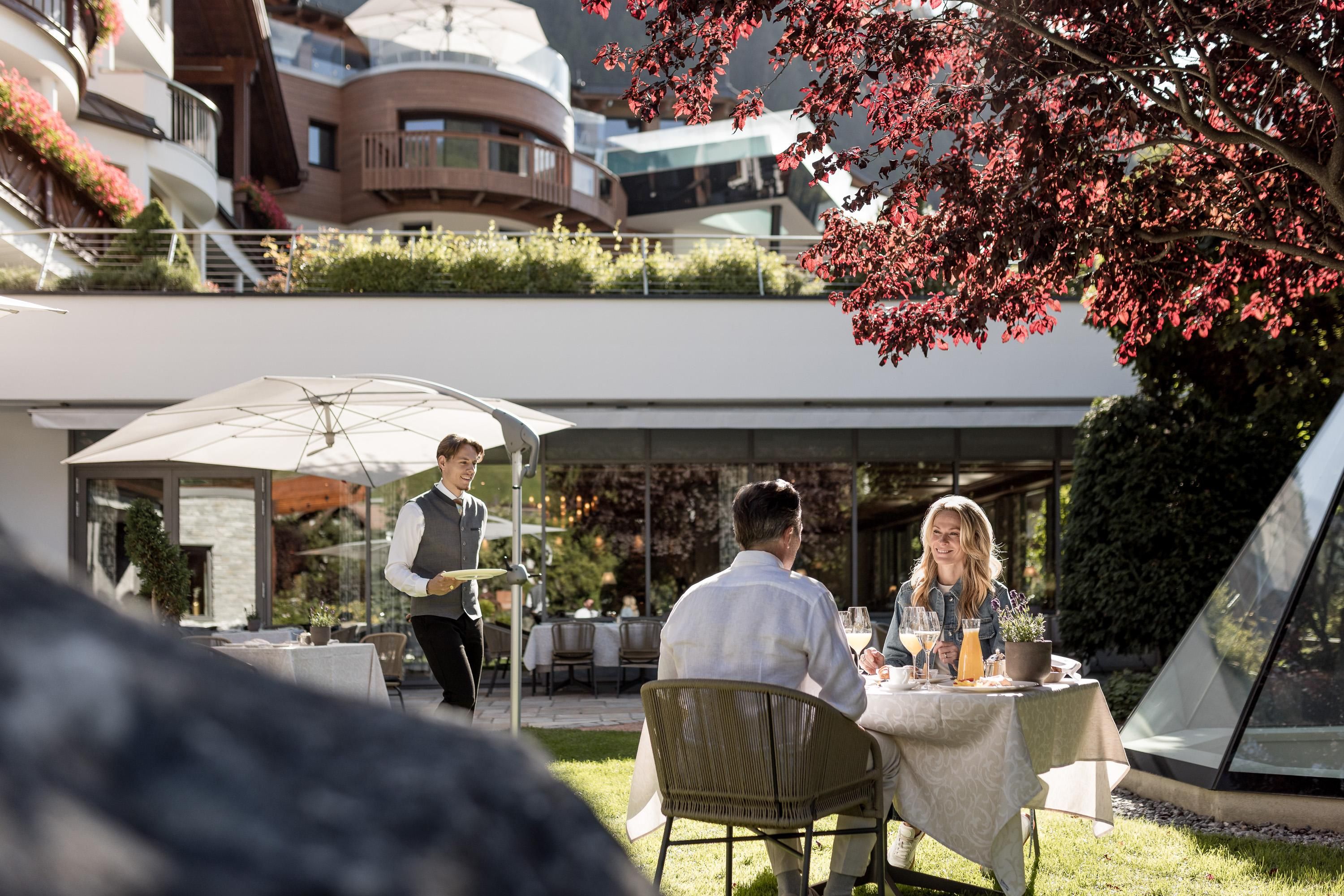 A beautiful outdoor restaurant with a table for two under a sunshade. A waiter serves drinks in a pleasant setting.