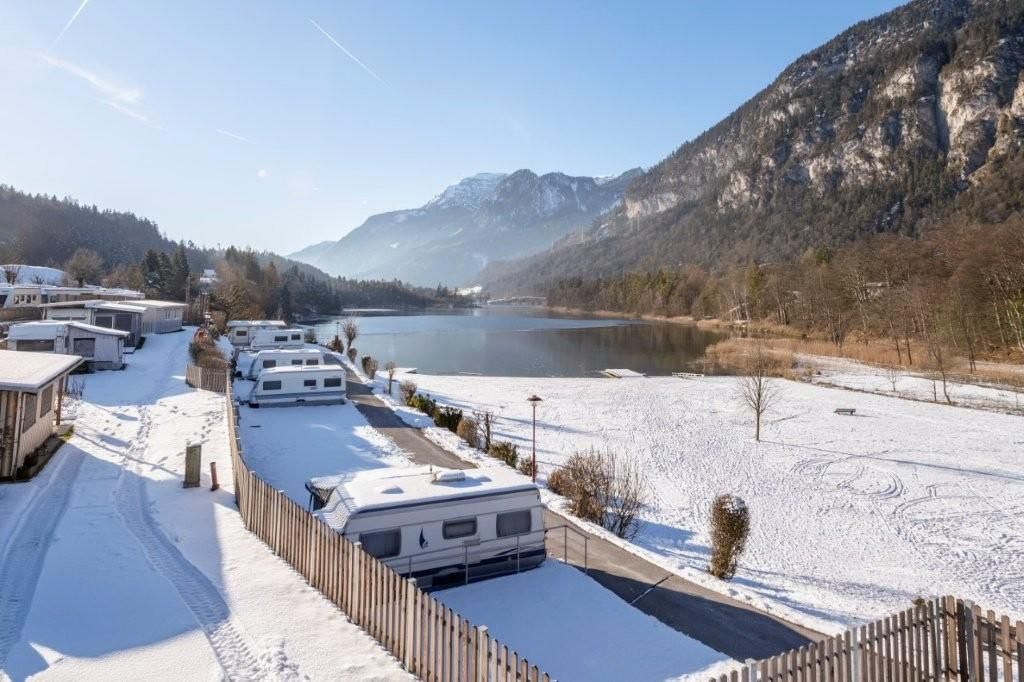 An idyllic winter landscape with snow-covered motorhomes and a serene lake. In the background, majestic mountains rise under a clear blue sky.