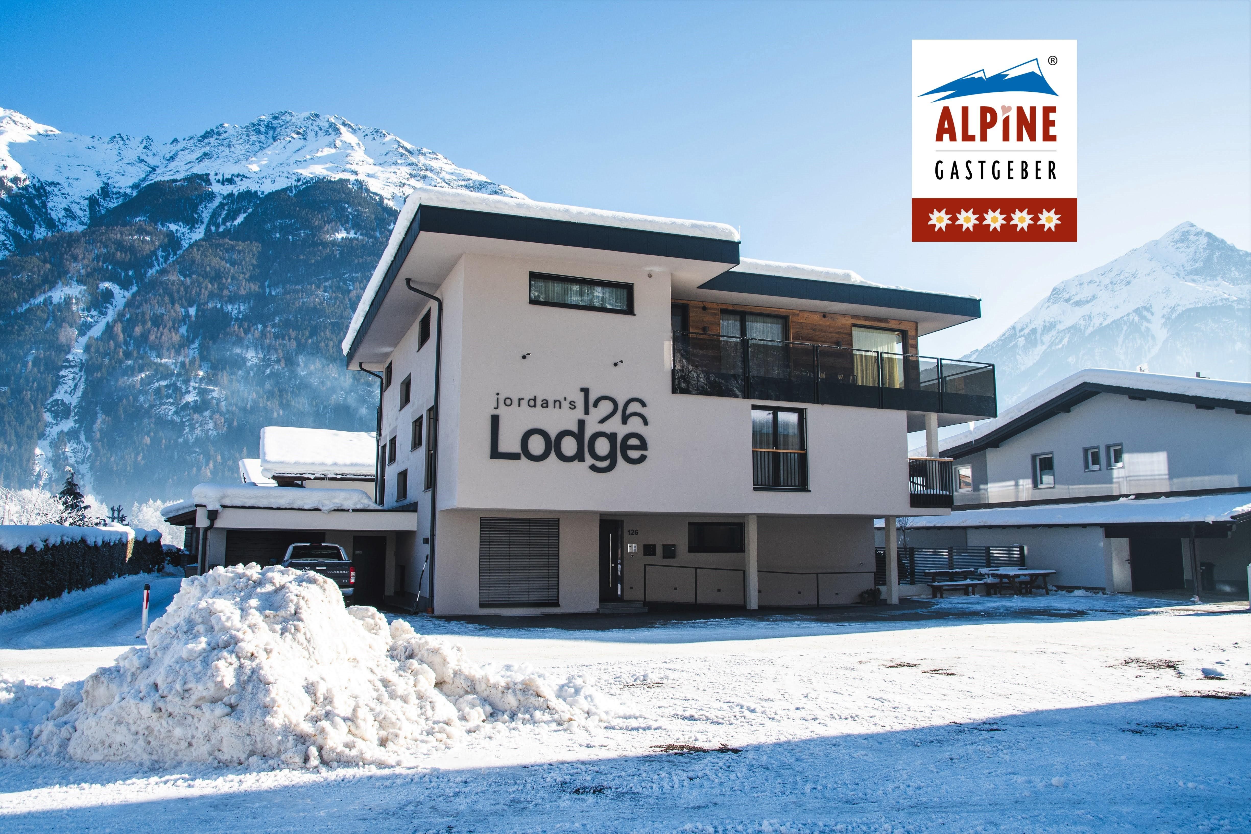A modern building named "Jordan's Lodge" in a snow-covered landscape. In the background, impressive snow-capped mountains can be seen.