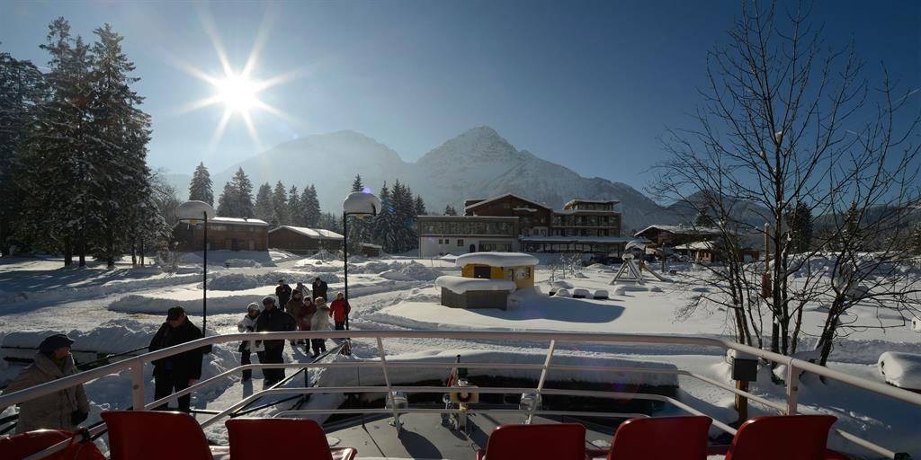 A snow-covered landscape with mountains in the background and the sun shining in the sky. People are strolling in the winter idyll.