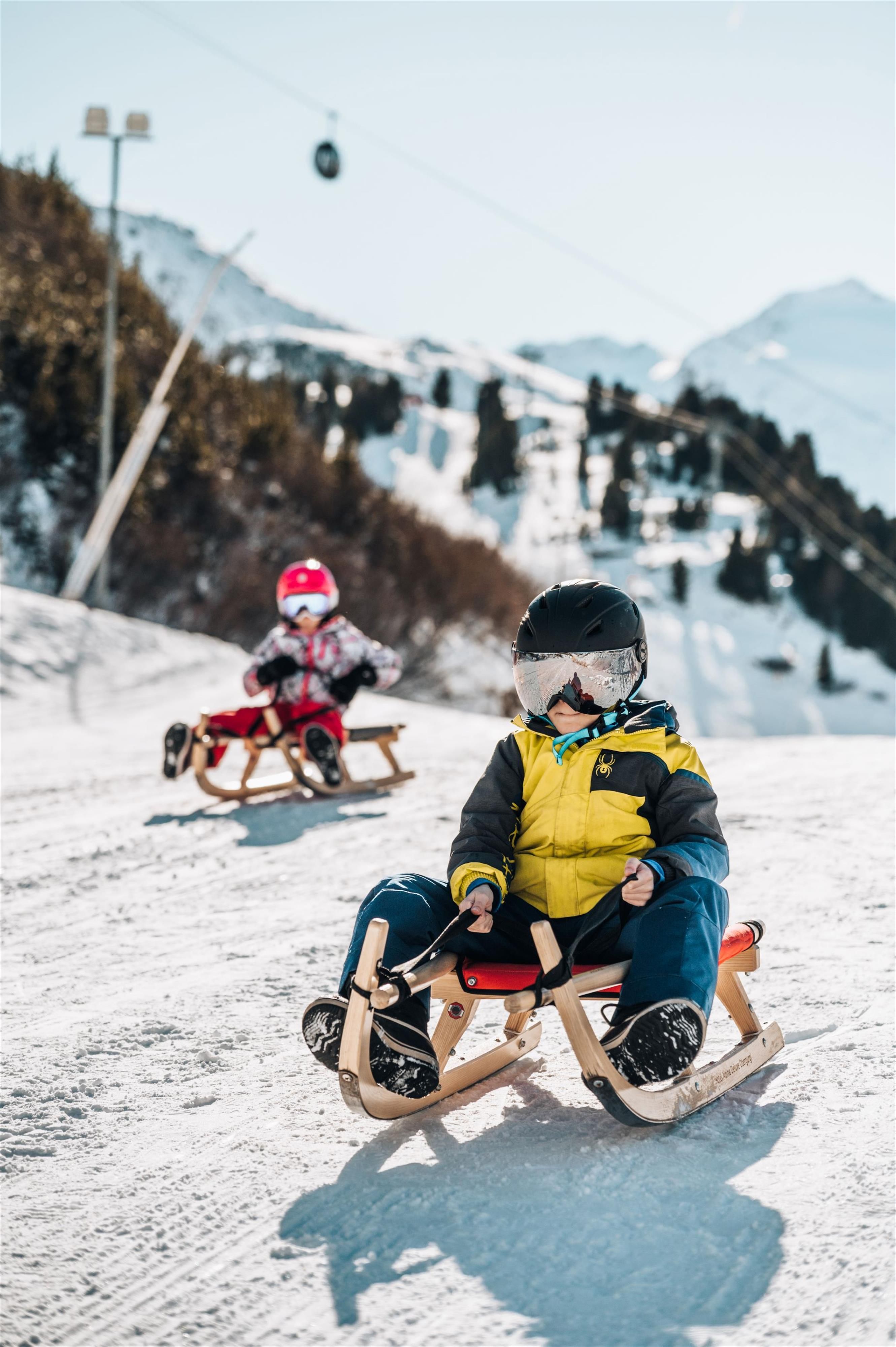 Two children are riding a sled down the snowy hill. The sun is shining and mountains can be seen in the background.
