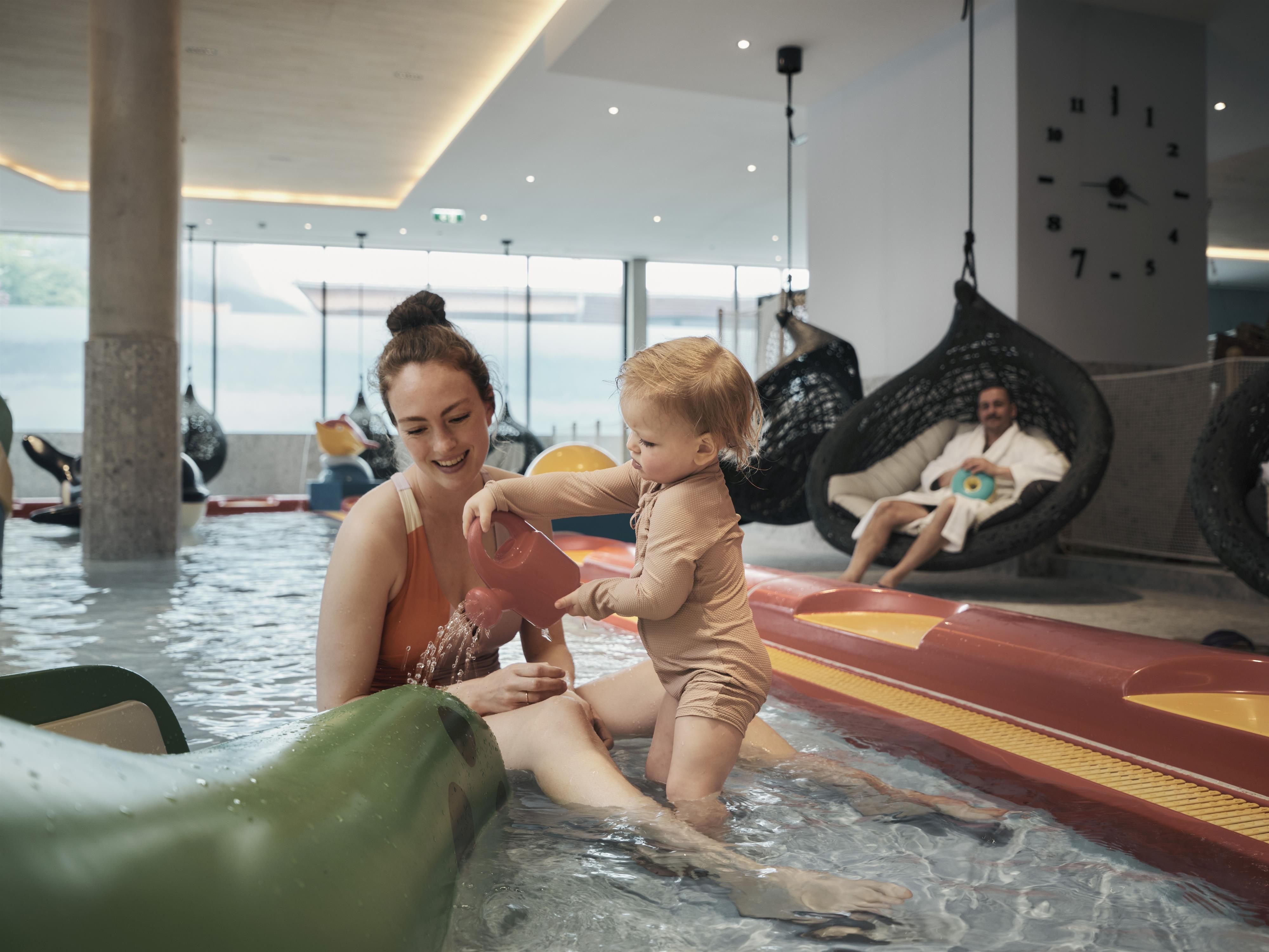A mother is playing with her child in the water of a swimming area. In the background, relaxing seating is visible.