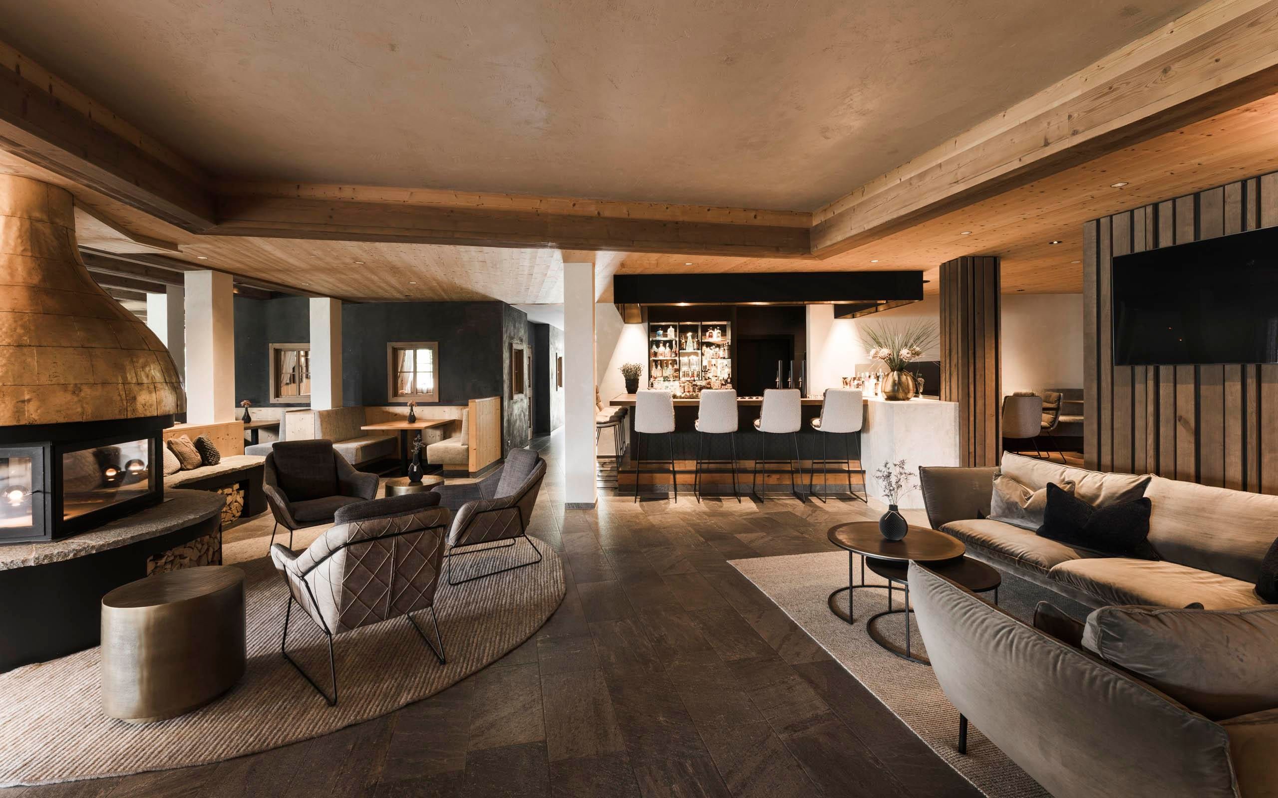 A modern lounge interior with cozy seating and an elegant fireplace. In the background, there is a stylish bar with stools.