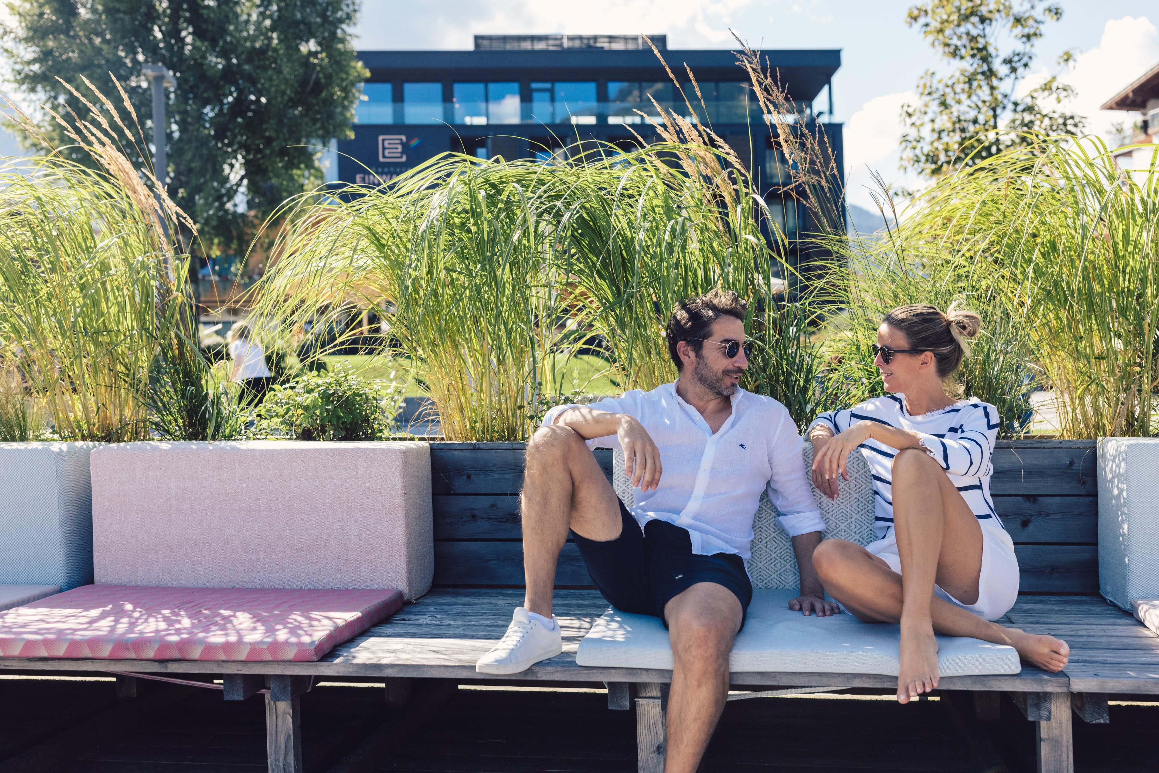 A couple relaxing on a bench surrounded by green plants. They are enjoying the sunny day in a modern environment.