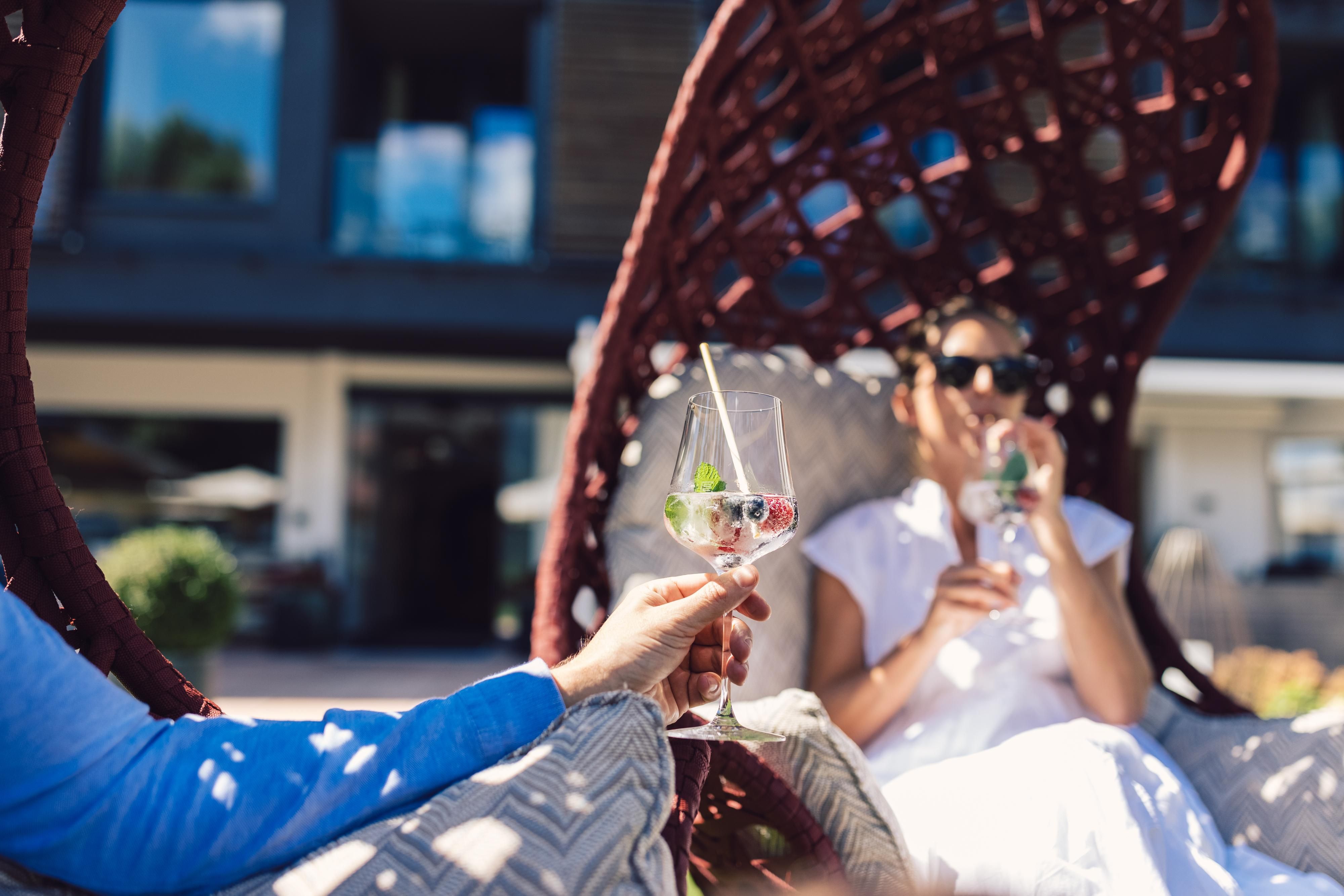 Two people are relaxing in a modern rocking chair and enjoying cocktails. The background features a beautiful ambiance with sun and plants.