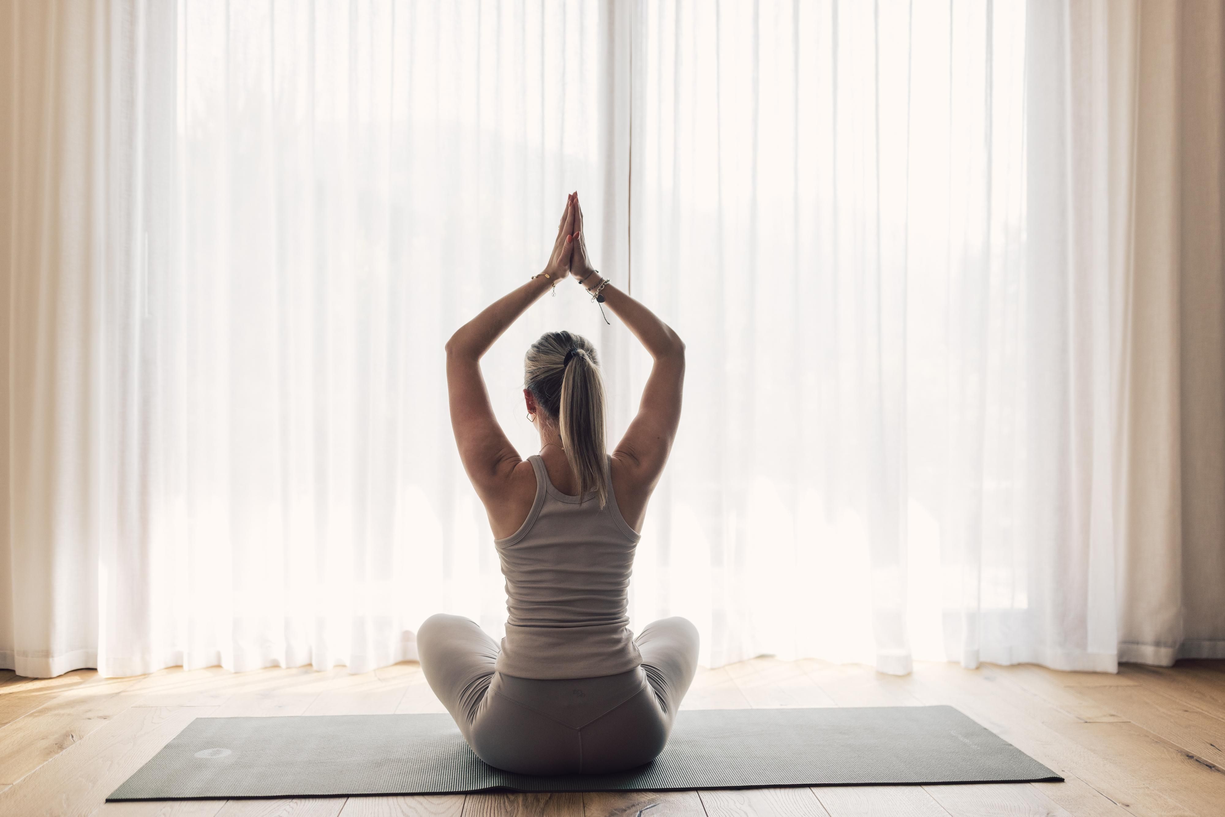 A person is sitting on a yoga mat and meditating. They have their hands clasped above their head and are looking forward into a bright room.