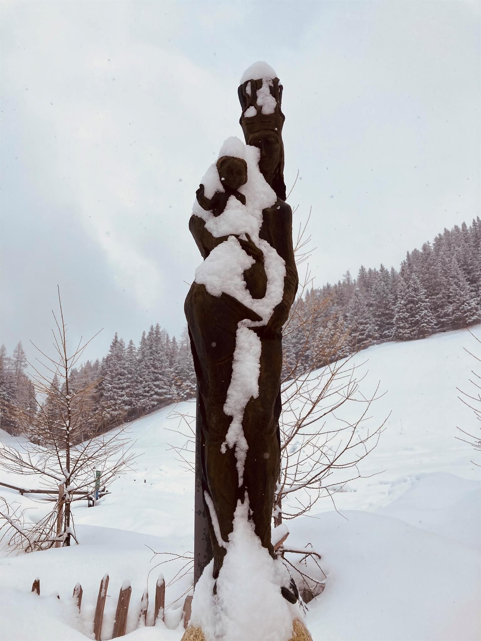 A wooden sculpture covered in snow. In the background, there are snowy trees and a wintry landscape.