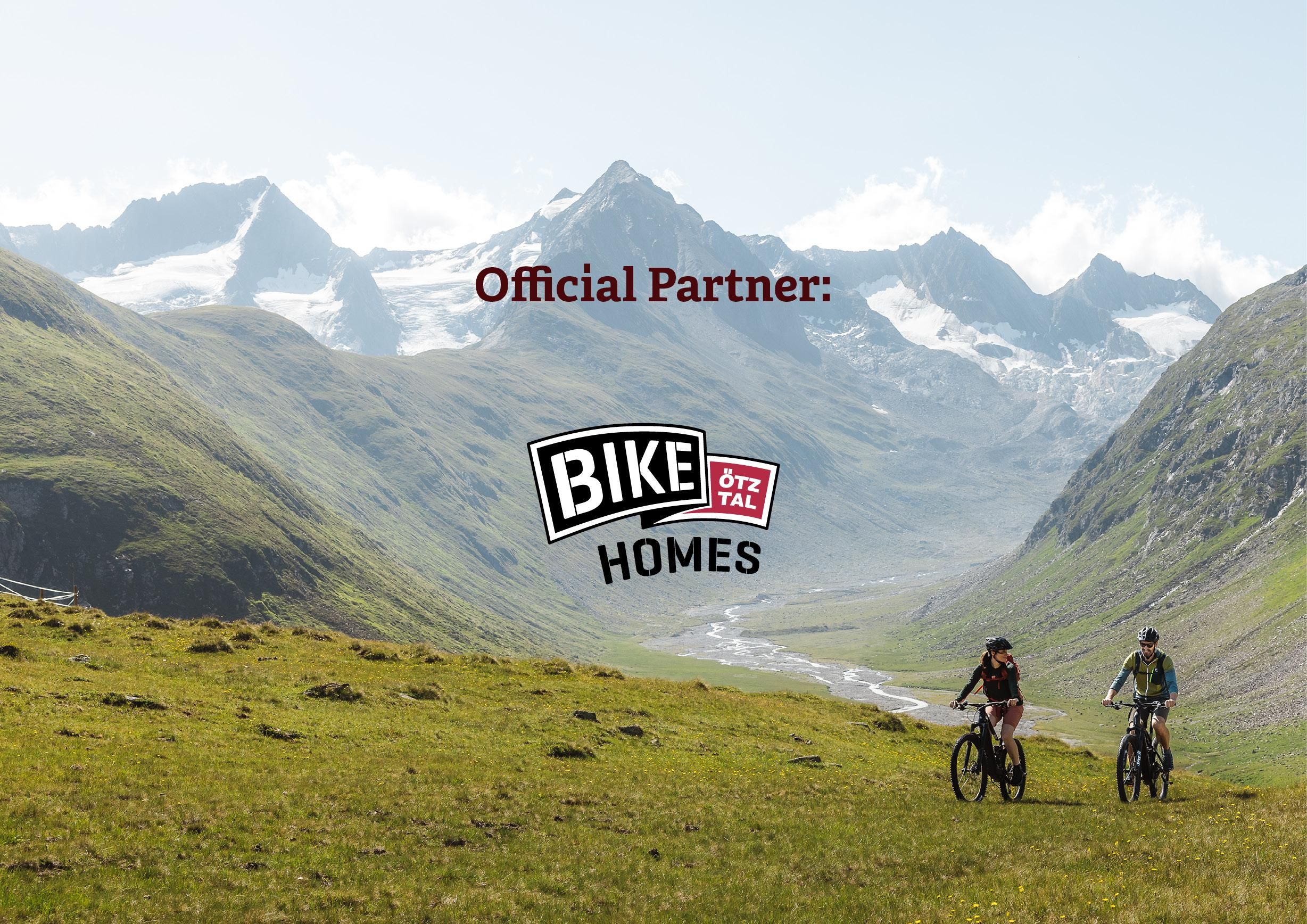 Two cyclists are exploring a picturesque mountain landscape with green meadows and impressive mountains in the background. The "Bike Homes" logo is visible at the bottom of the image.