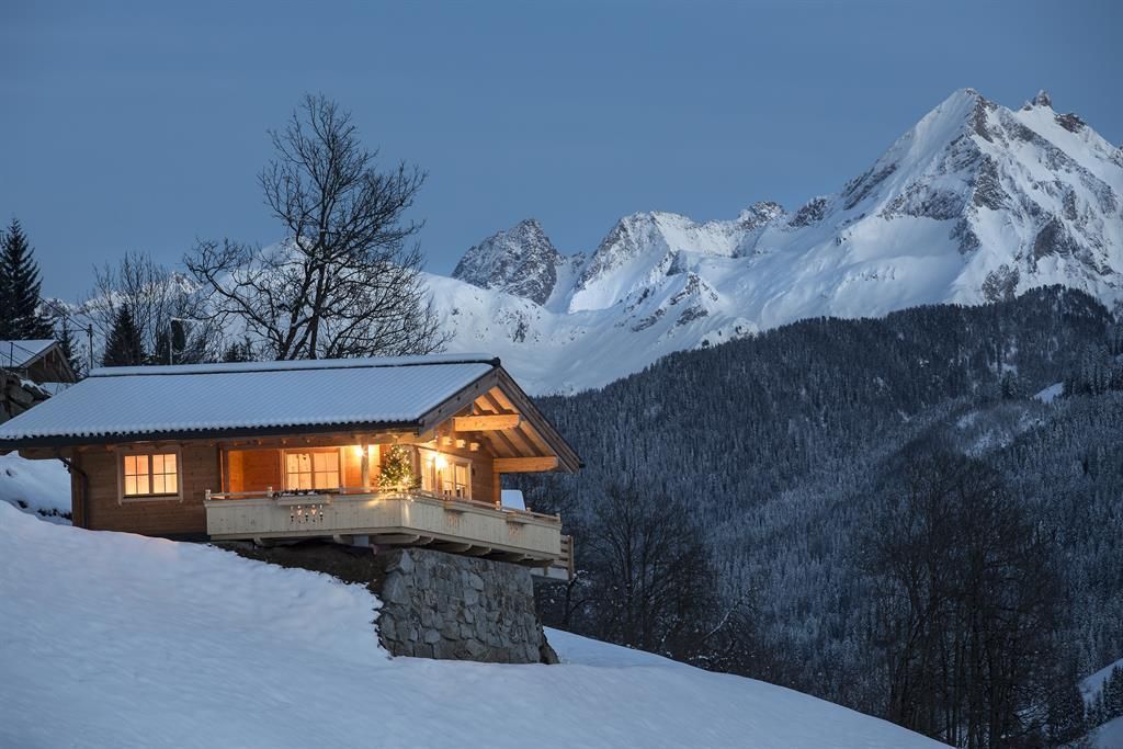 A cozy chalet in a snowy landscape, surrounded by mountains. Warm light radiates from the windows at dusk.