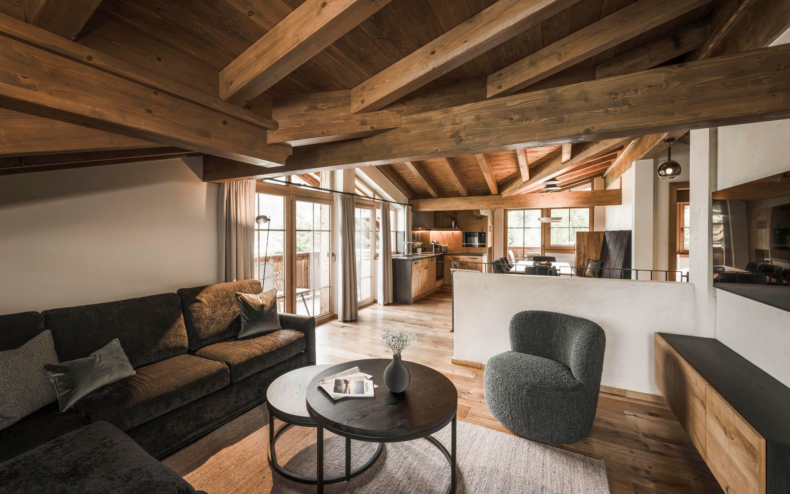 A cozy living space with a wooden ceiling and large windows. The decor is modern, featuring a sofa, a round table, and chairs.