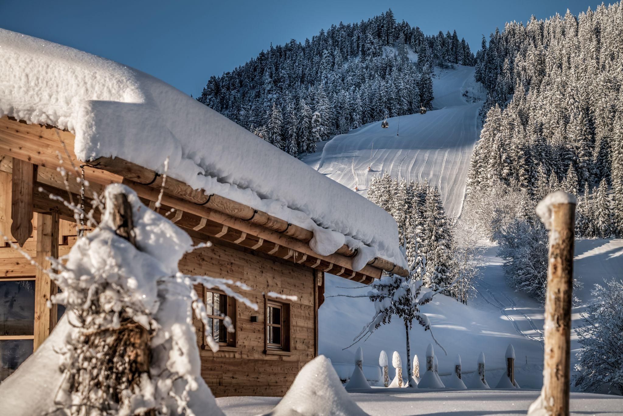 A cozy chalet in the snow with a thick layer of snow on the roof. In the background, you can see snow-covered mountains and trees.