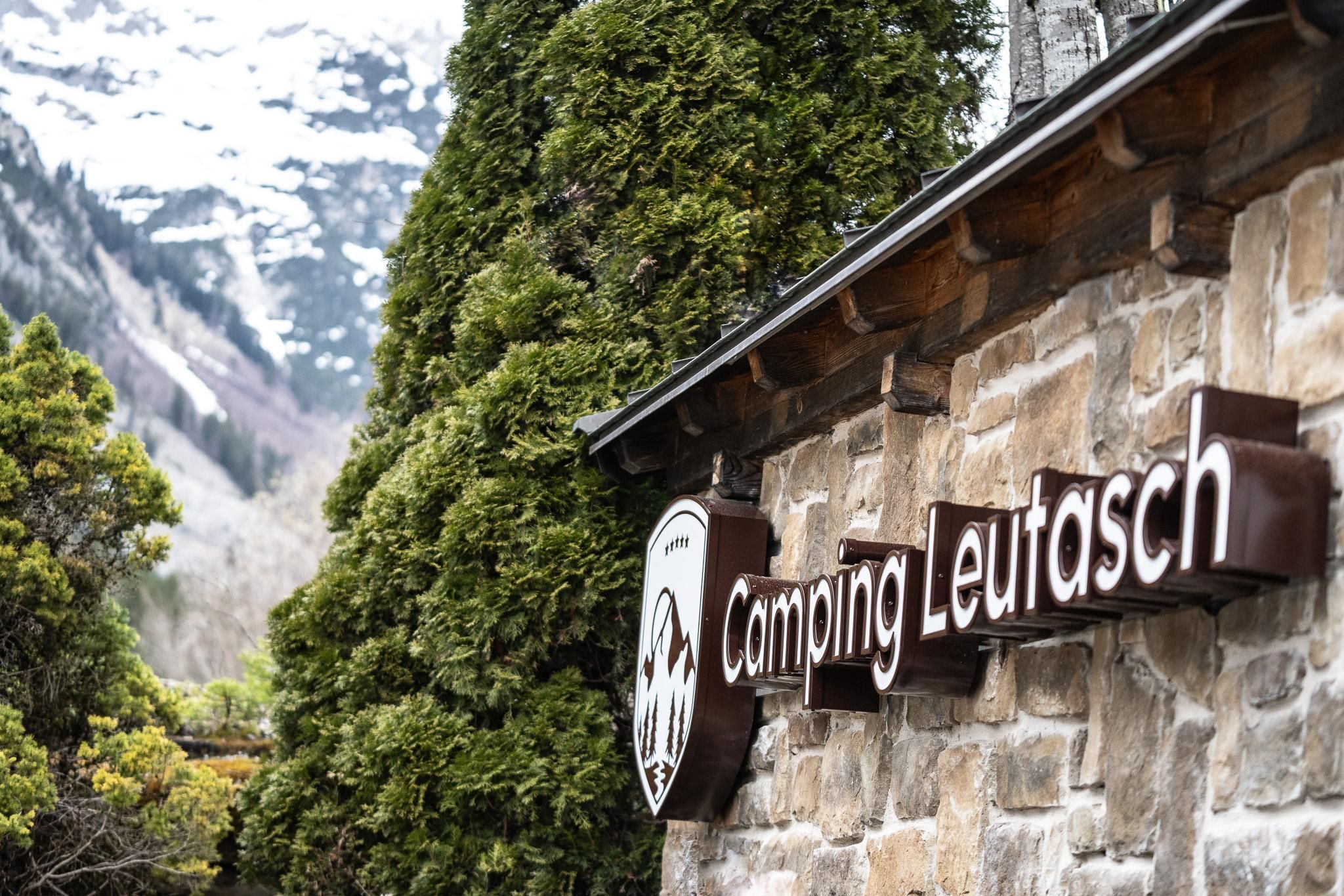 A wooden sign with the inscription "Camping Leutasch" in a natural setting. Mountains and trees can be seen in the background.