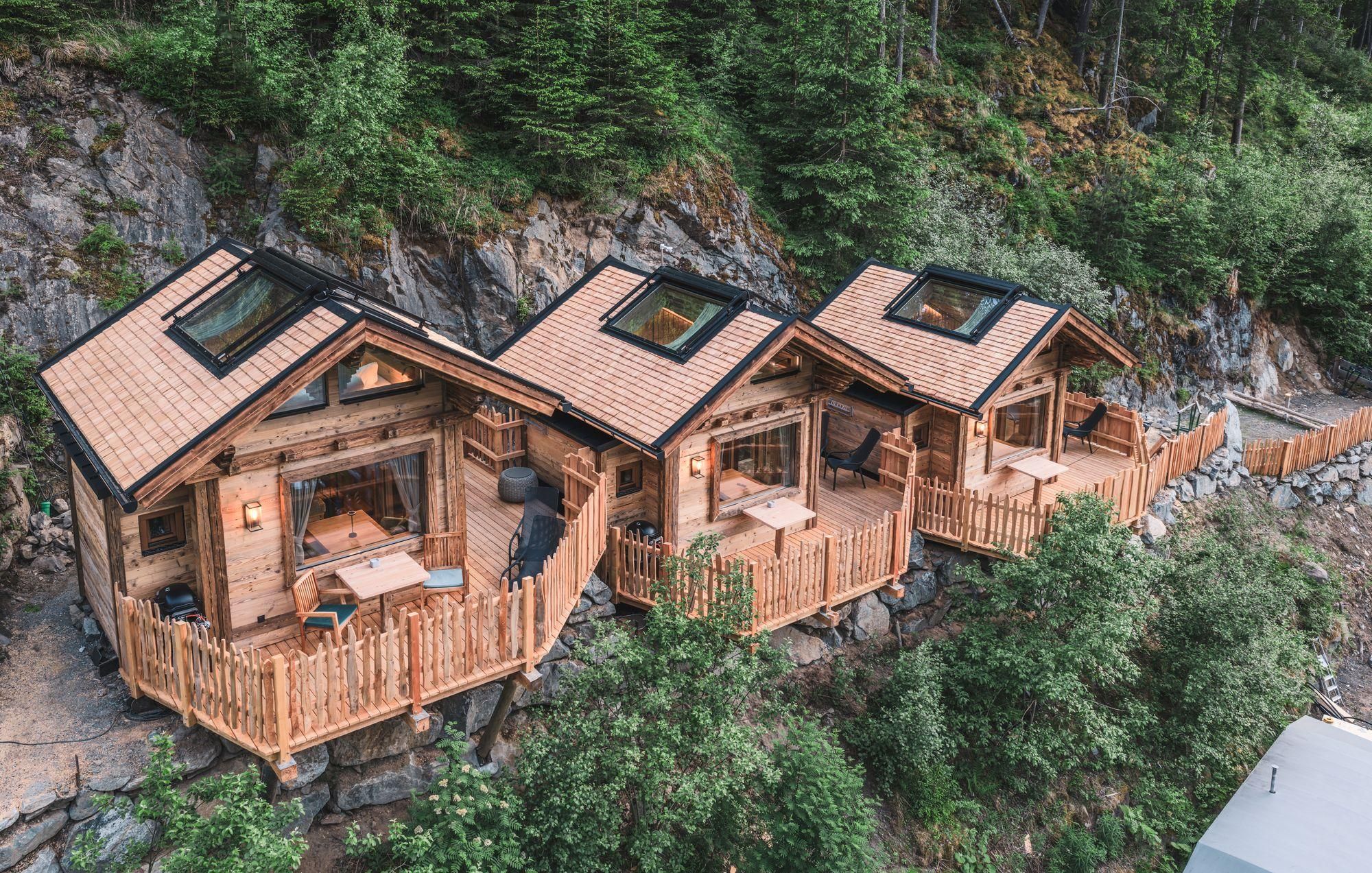 Three wooden cottages stand in nature, surrounded by trees. Each house has a porch with a view of the surroundings.