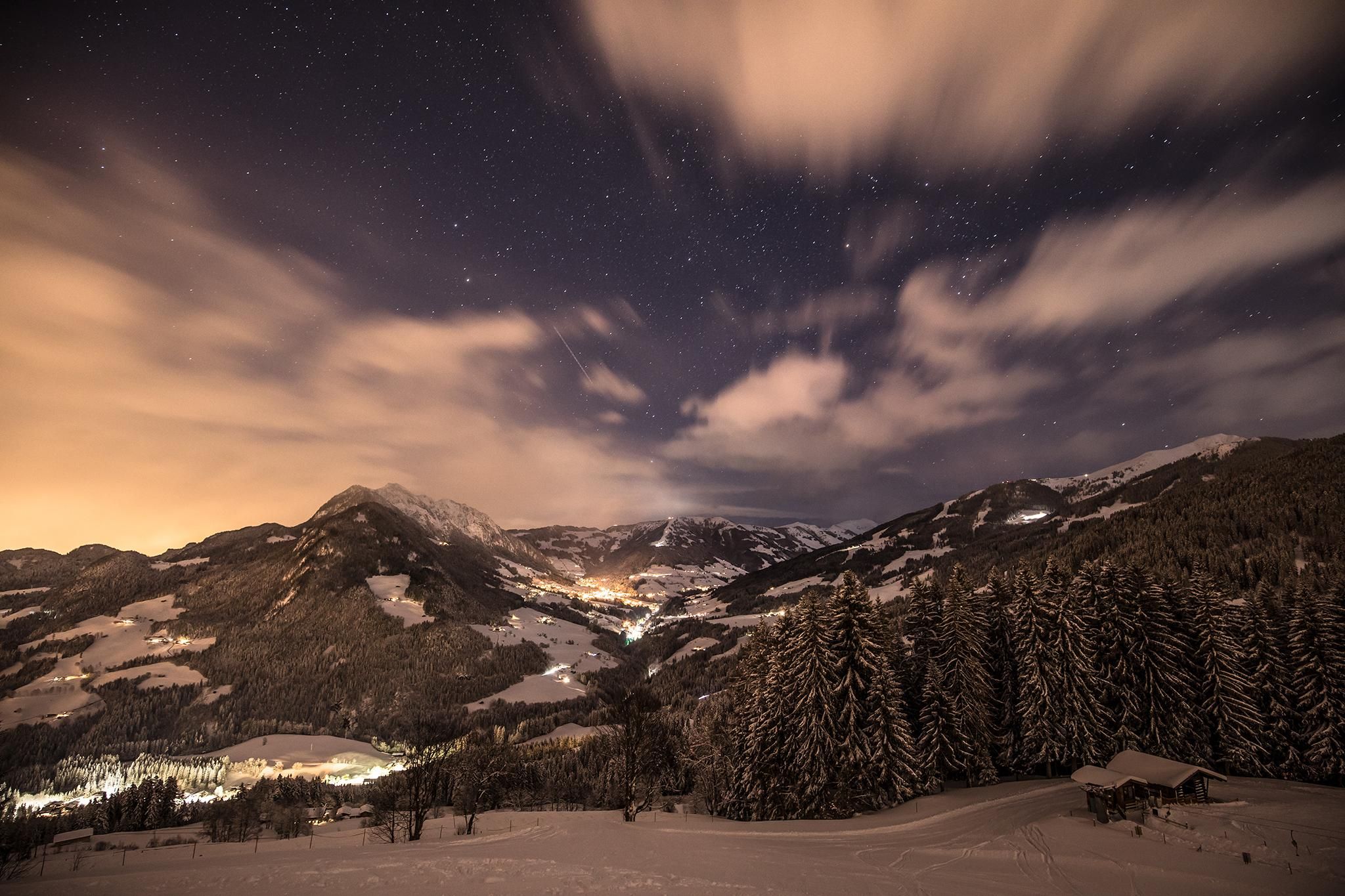 A winter mountain landscape under a clear starry sky. The snow-covered hills and trees reflect the faint light of the city in the background.