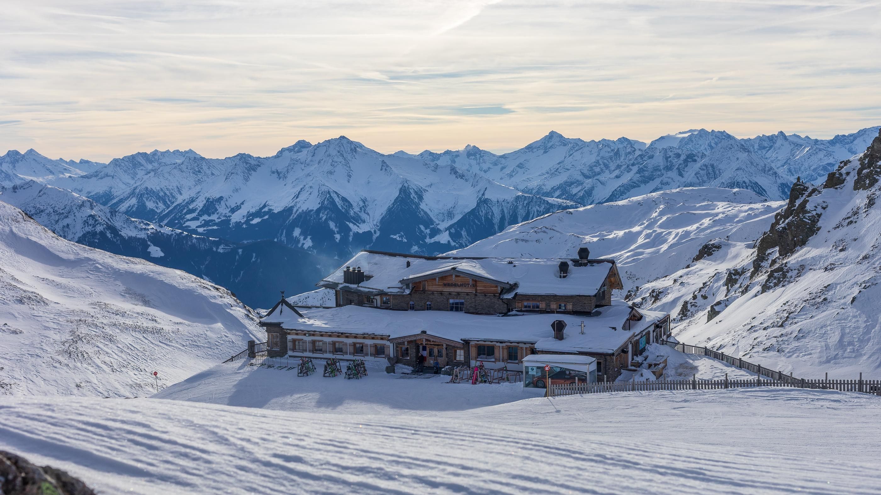 A snow-covered mountain area with a rustic building. The surrounding mountains are majestic and the sky is overcast.