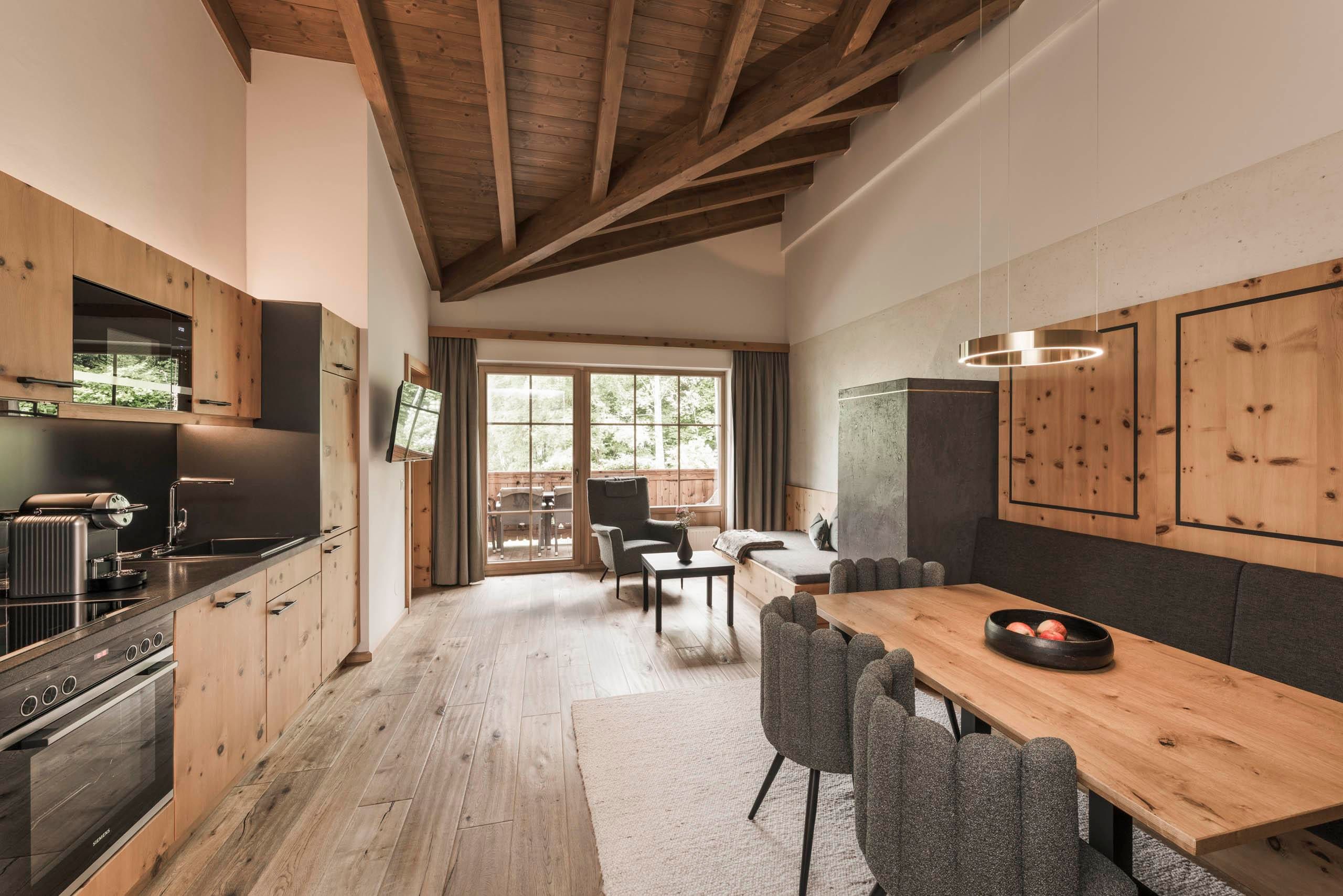 A modern living room with an open kitchen and wooden elements. Large windows offer a view of a balcony and ensure plenty of light.