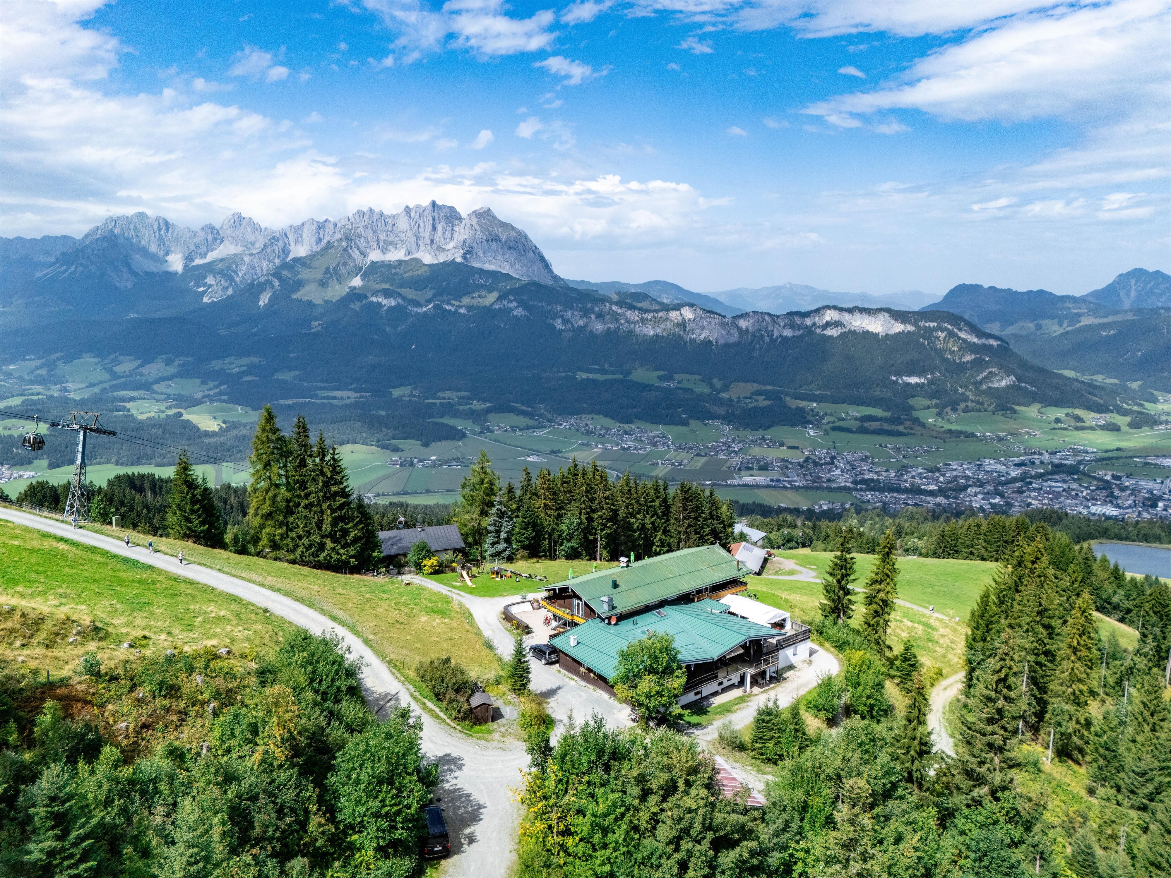 A picturesque mountain landscape with high peaks and a green meadow. In the foreground stands a cozy mountain hut surrounded by fir trees.