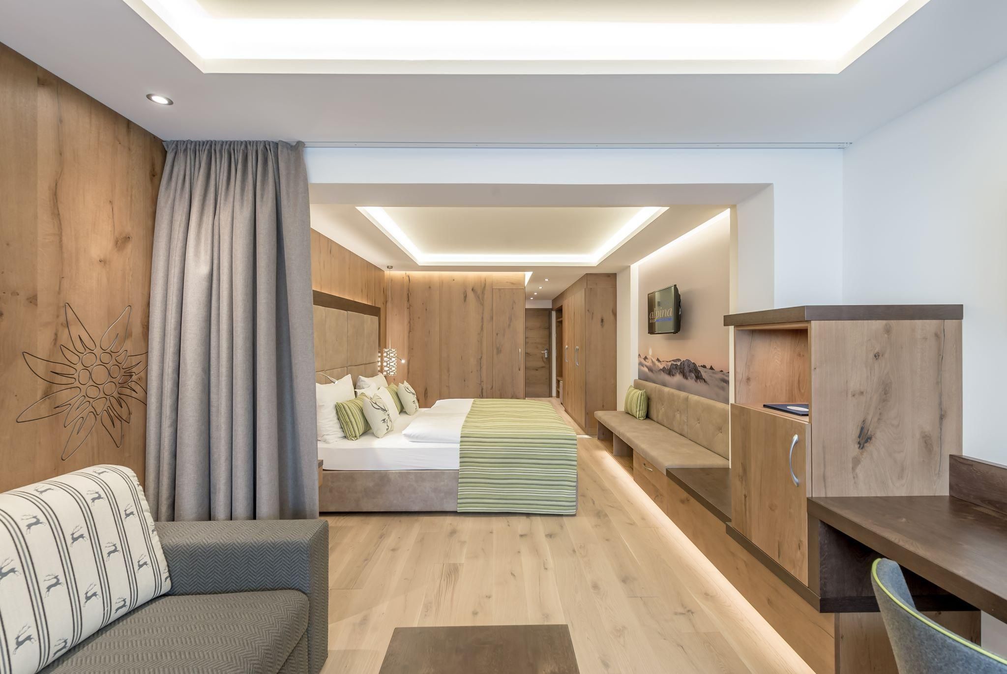 A modern hotel room with a double bed, a sofa, and a wooden wardrobe. The warm colors and soft light create a cozy atmosphere.