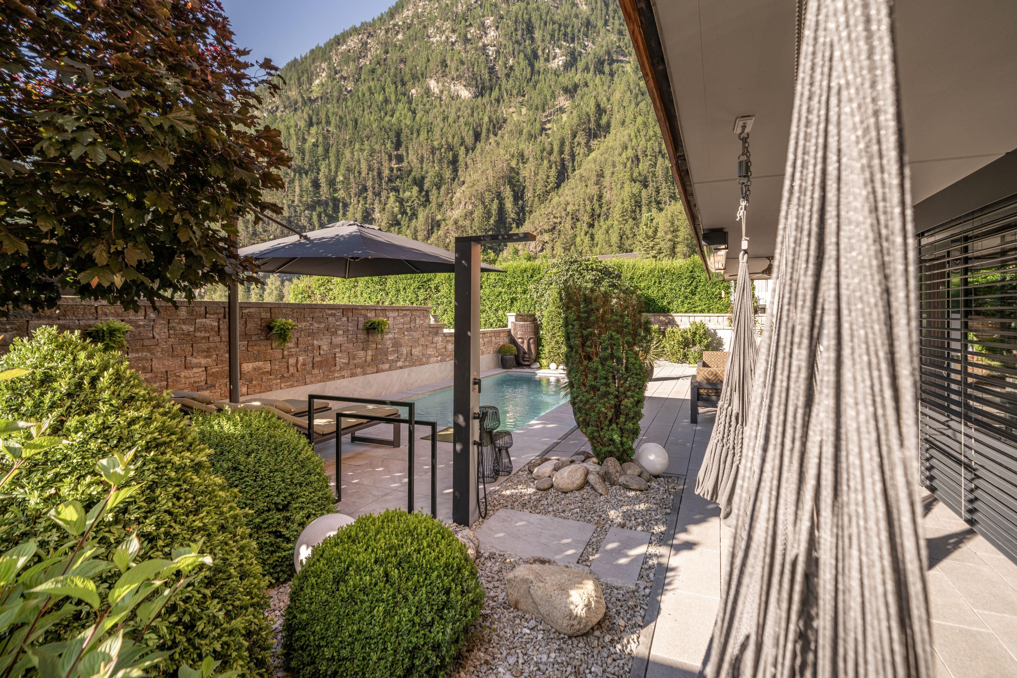 A beautiful garden with a pool and well-maintained green bushes. In the background, one can see tall mountains and a blue sky.