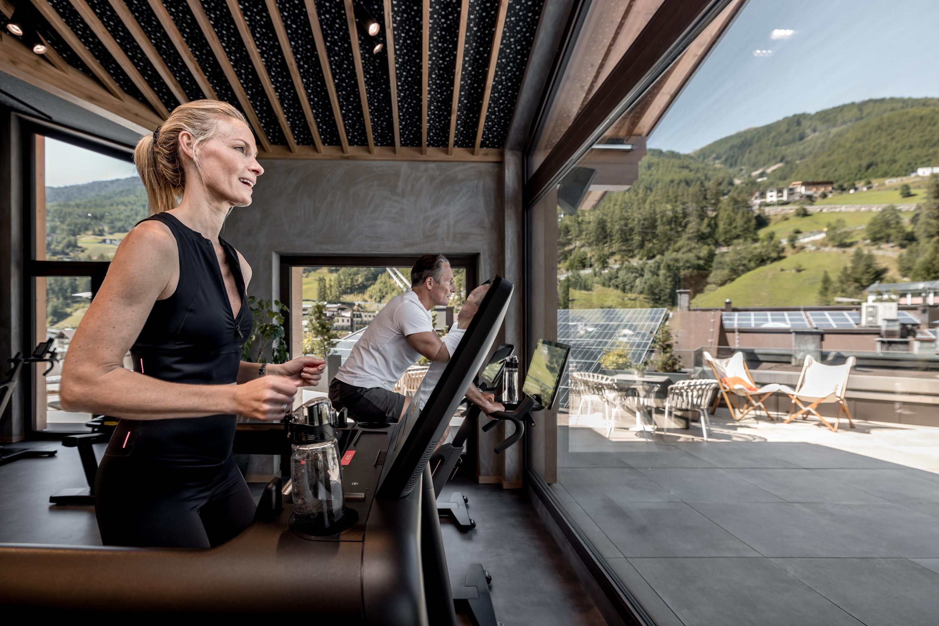 A woman is exercising on a treadmill in a modern gym with large windows. In the background, green hills and a sun terrace are visible.